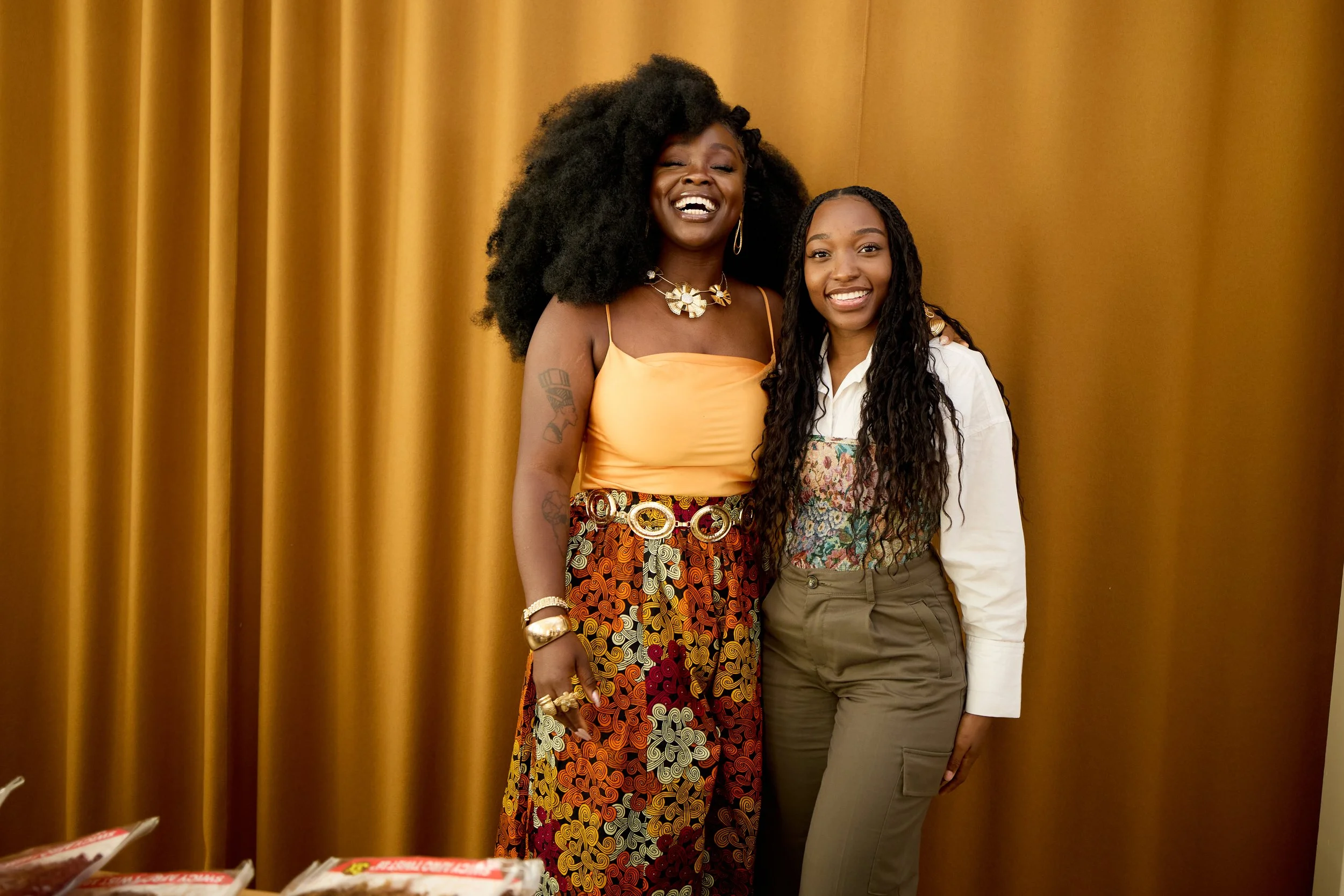 Two women standing in front of a gold curtain, smiling at the camera. The woman on the left has big curly hair and is wearing a yellow top and colorful patterned skirt with jewelry. The woman on the right has long curly hair and is wearing a white sh