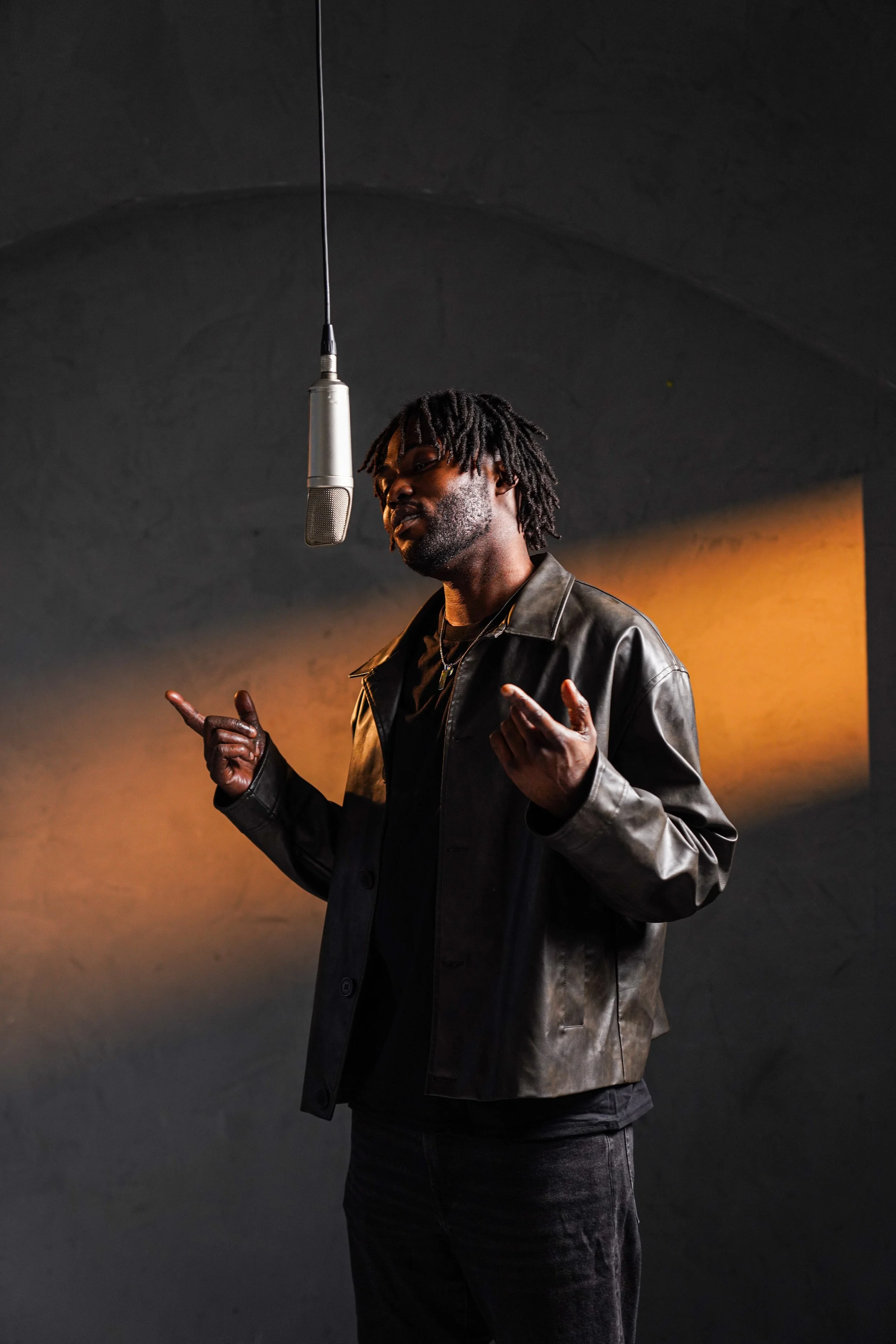 A man with dreadlocks and a beard standing in front of a dark wall, wearing a black leather jacket and speaking into a microphone hanging from the ceiling.