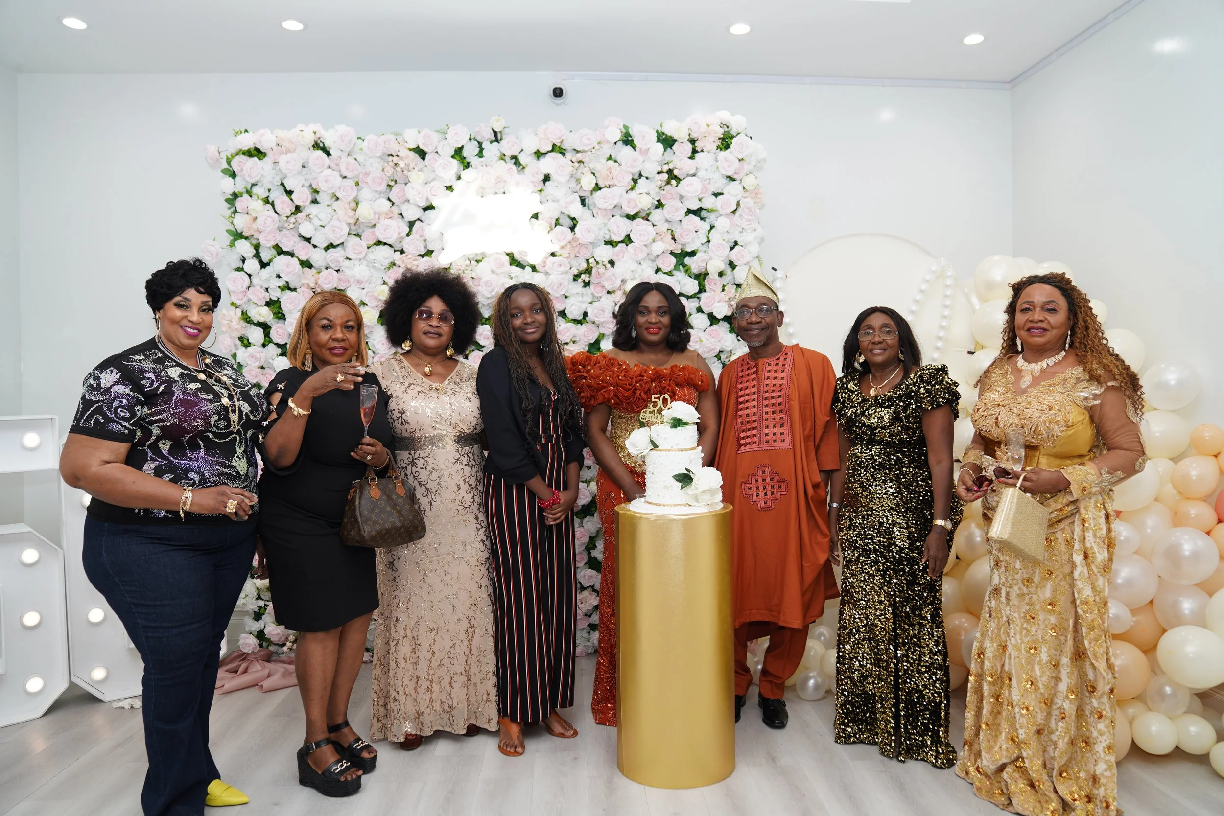Group of nine people standing in front of a floral backdrop, celebrating a 50th birthday with a cake, wearing formal and traditional attire. One person is wearing a party hat, and some are holding glasses.