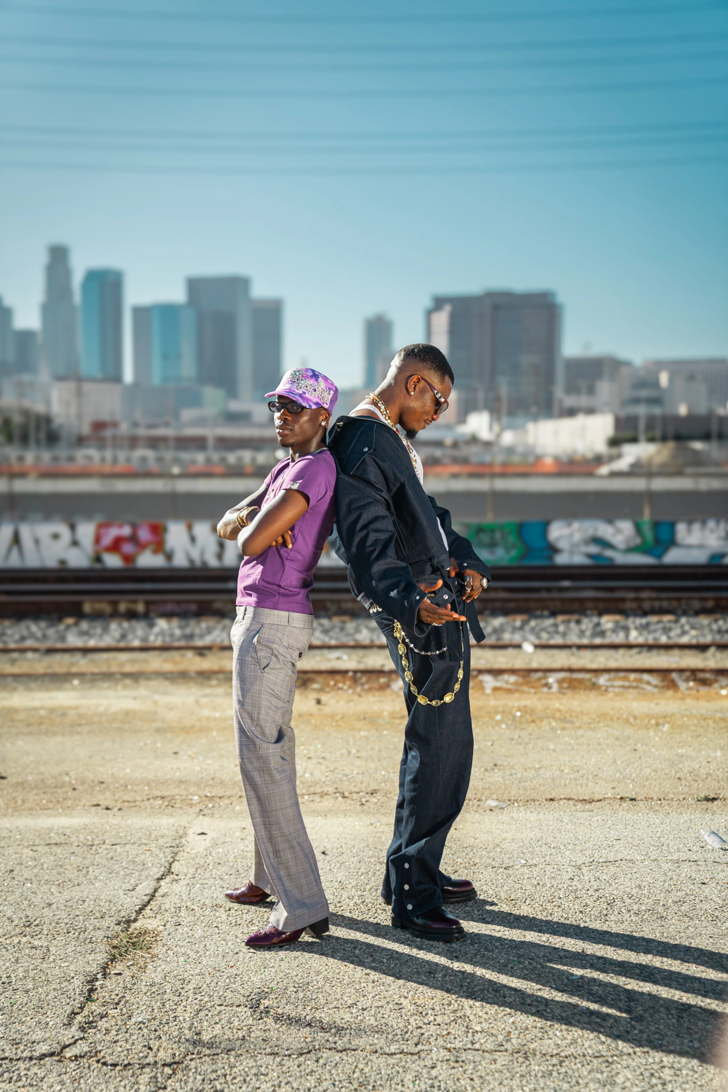 Two young men standing back-to-back on an urban street near train tracks with a city skyline in the background, wearing stylish casual outfits and sunglasses.