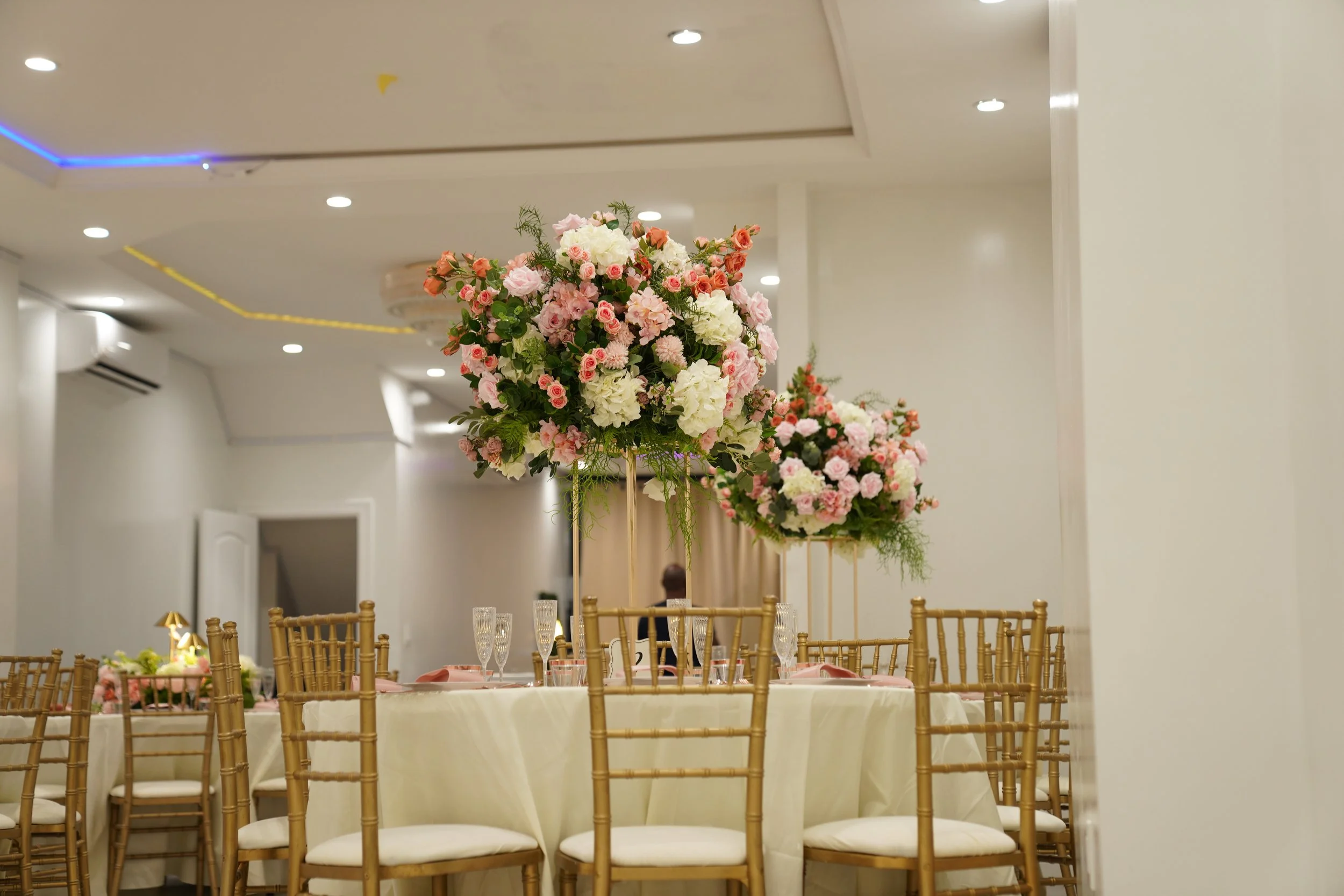 Elegant event table with gold chairs and tall floral centerpieces in a well-lit banquet hall.