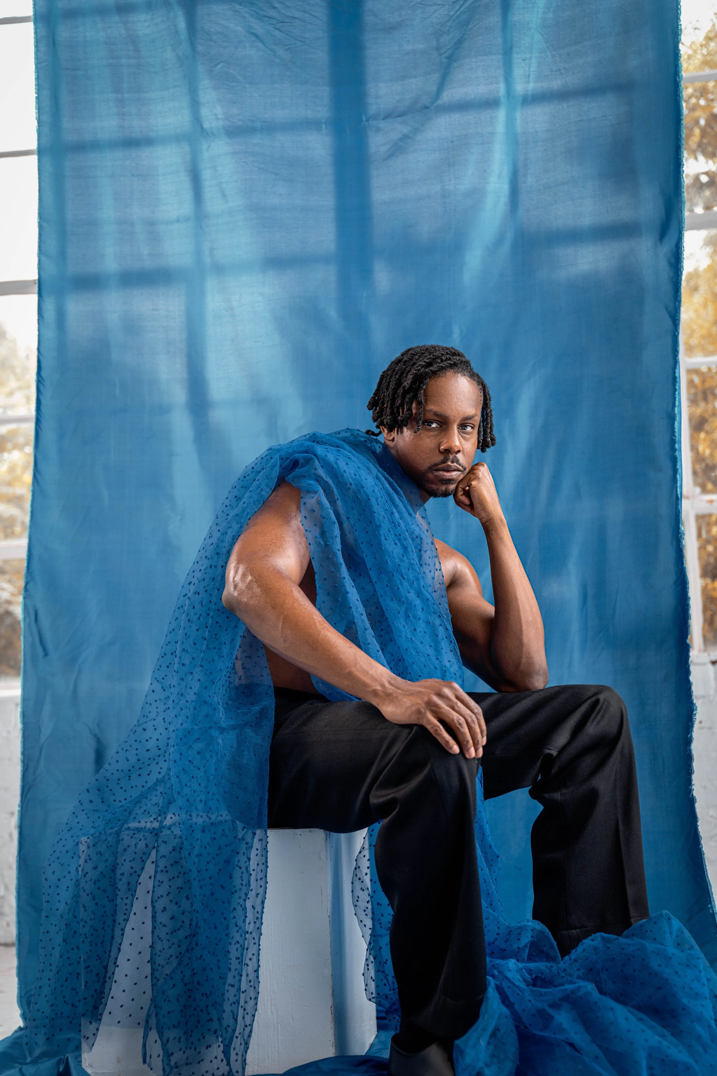 A man sitting on a white block, dressed in black pants with a blue sheer fabric draped around his shoulders, in front of a blue backdrop, with trees visible through the window behind.