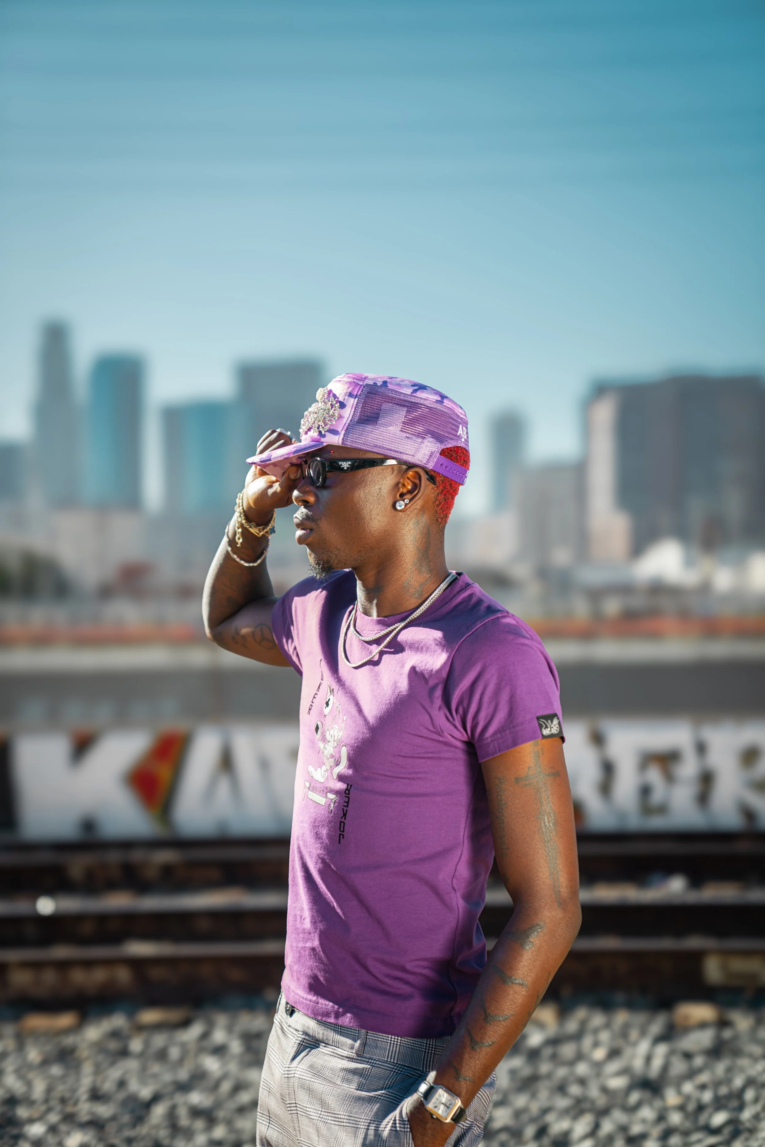 Young man with colorful hair wearing purple sunglasses, a purple t-shirt, and a purple cap, standing on train tracks with a city skyline in the background.