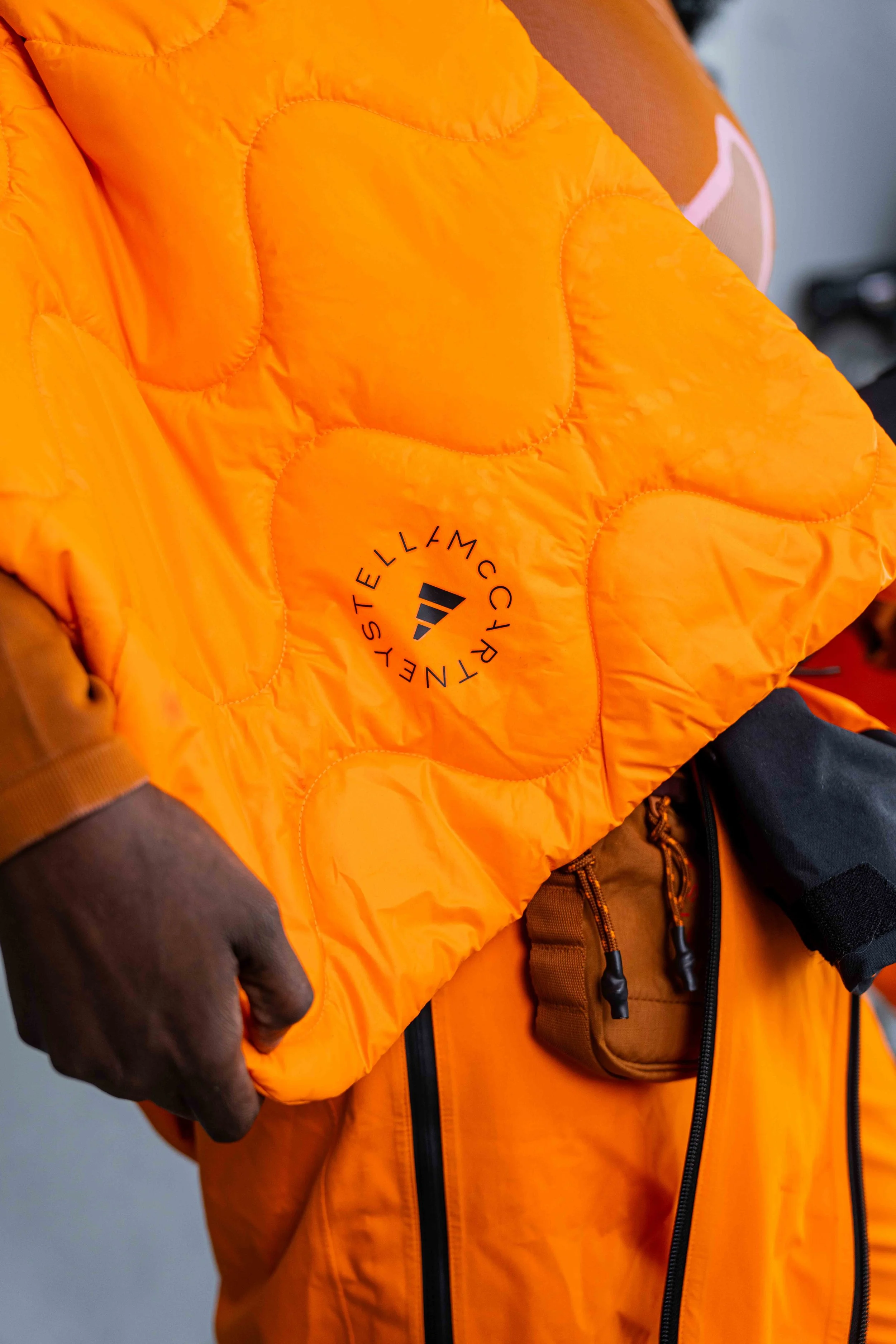 Close-up of a bright orange puffer jacket with a circular logo reading 'WELLSTYLEDWELL' and a small geometric emblem in the center.