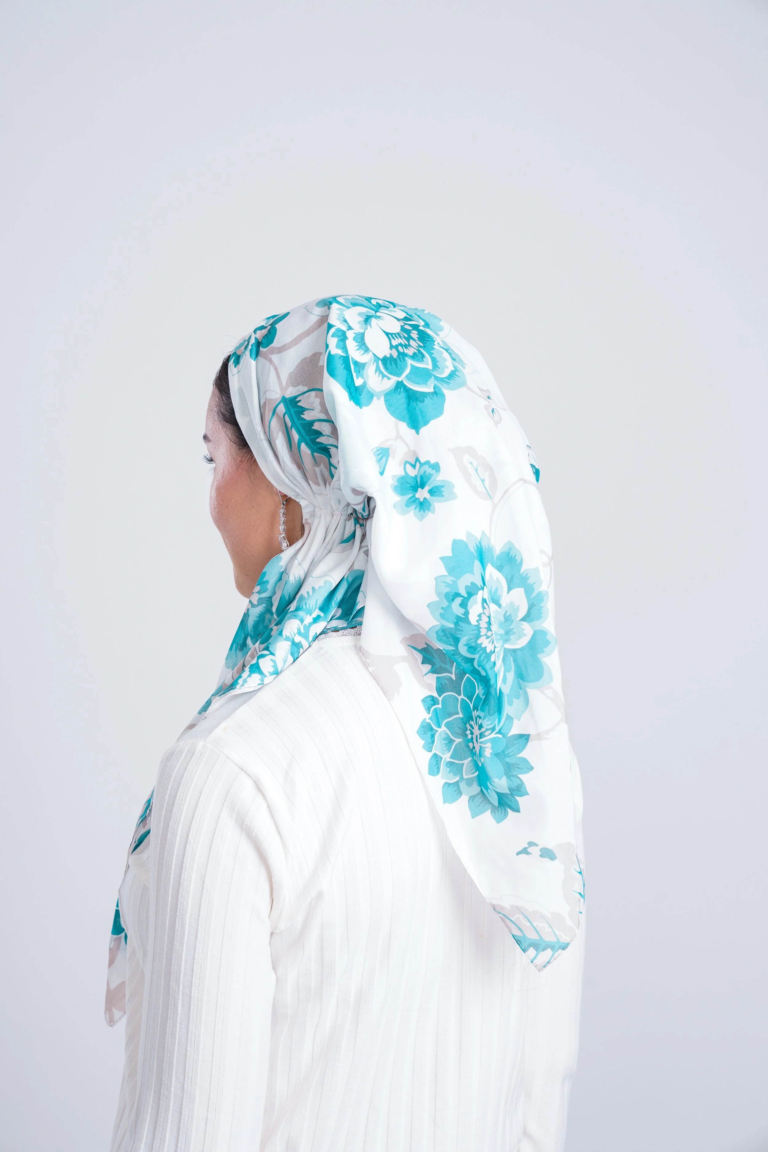 Profile of a woman wearing a white headscarf with blue floral patterns and a white ribbed top, against a plain light background.