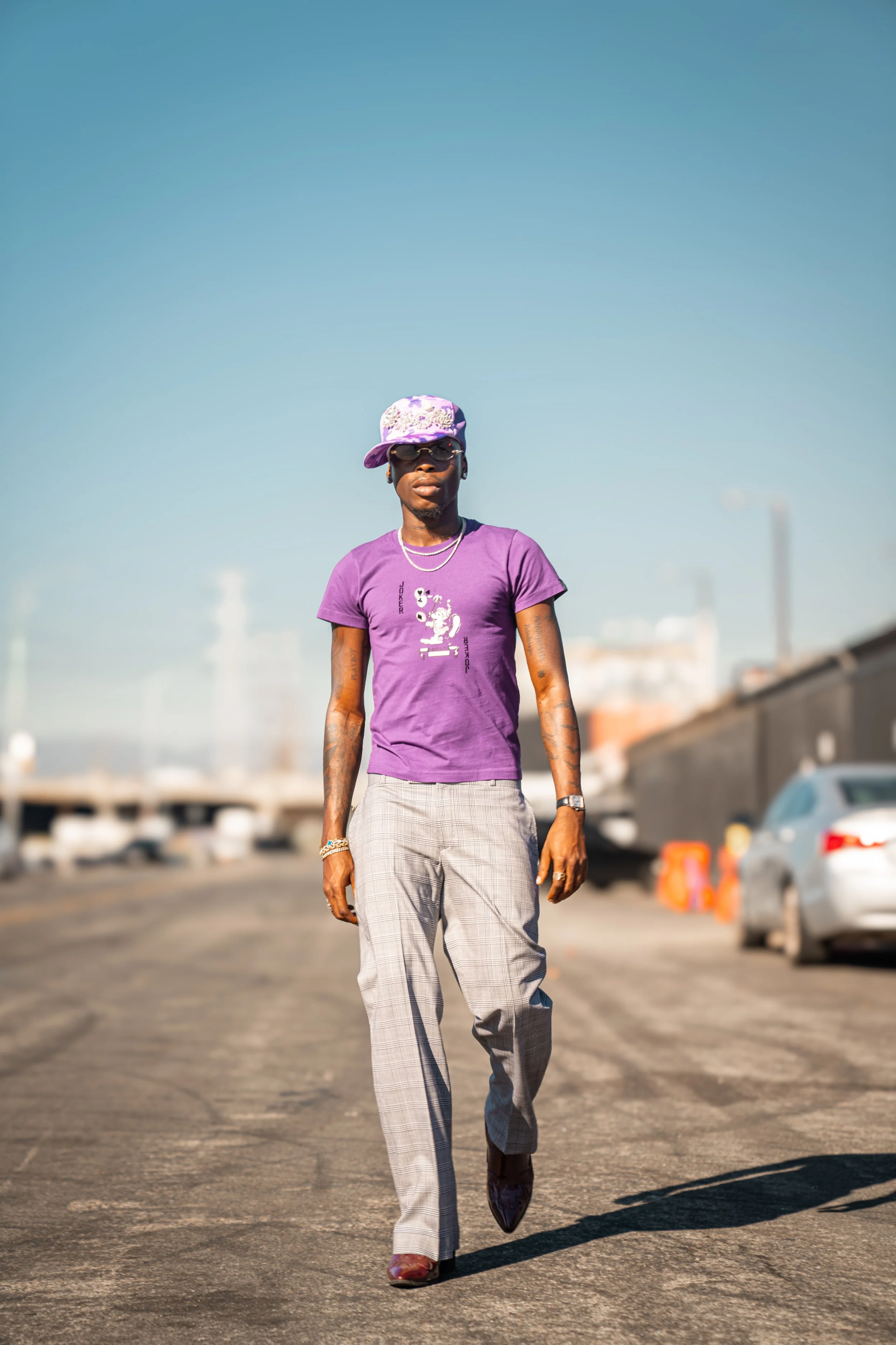 A stylish man walking down an urban street under a clear blue sky, wearing a purple t-shirt, plaid pants, a patterned purple hat, sunglasses, and accessories.