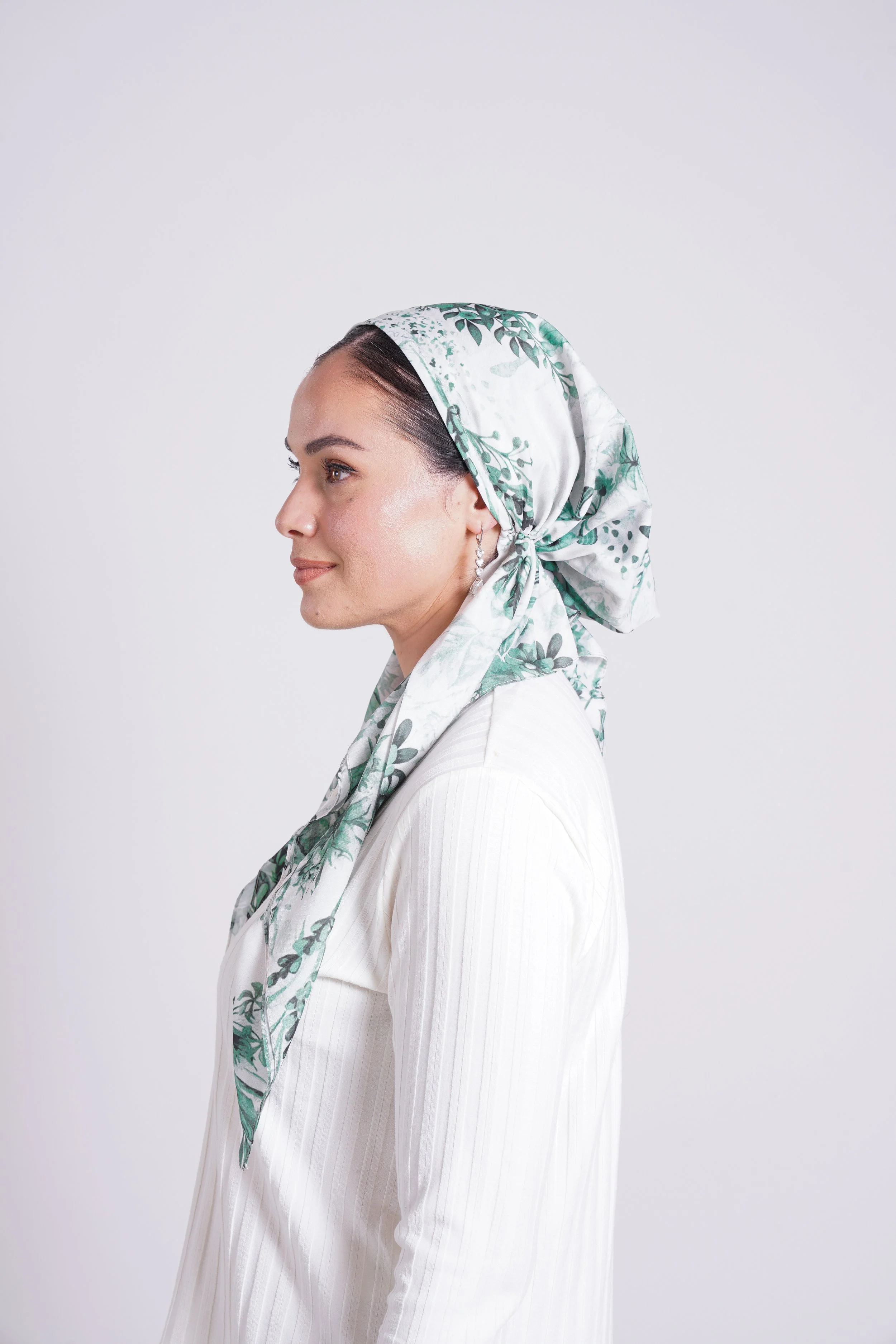 Side profile of a woman wearing a white shirt and a white headscarf with green leaf patterns, standing against a plain light background.