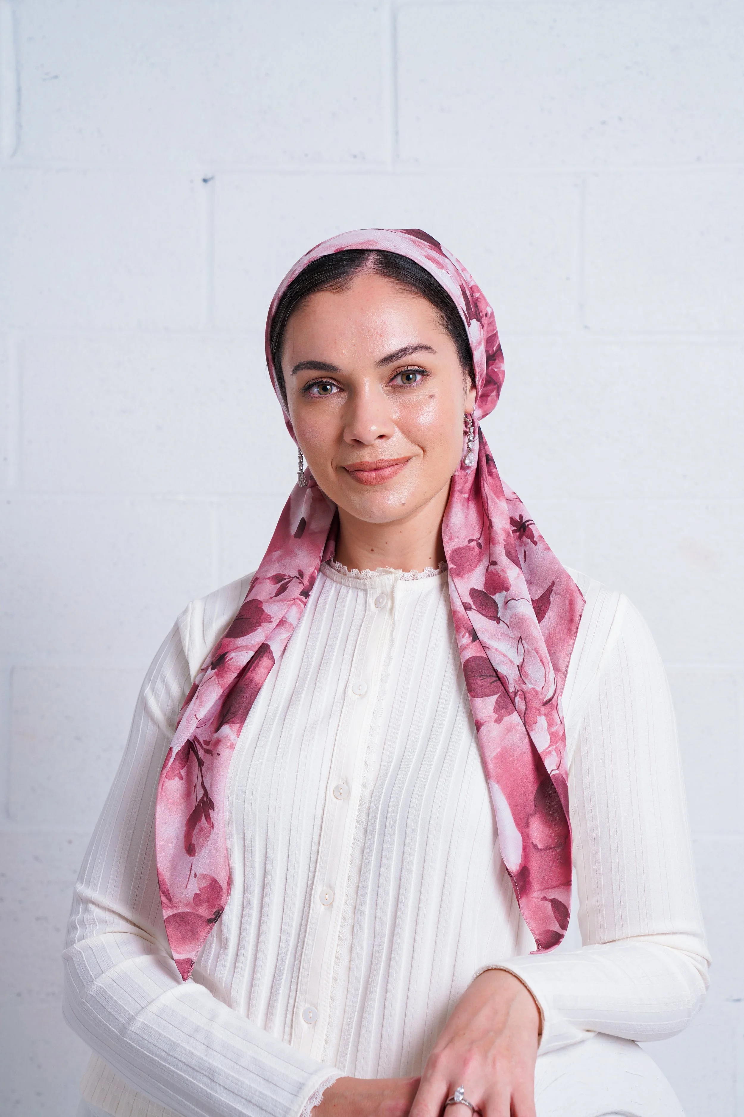 A woman with dark hair wearing a pink floral headscarf, earrings, a white blouse with vertical stripes, and a ring, smiling and standing against a white brick wall background.