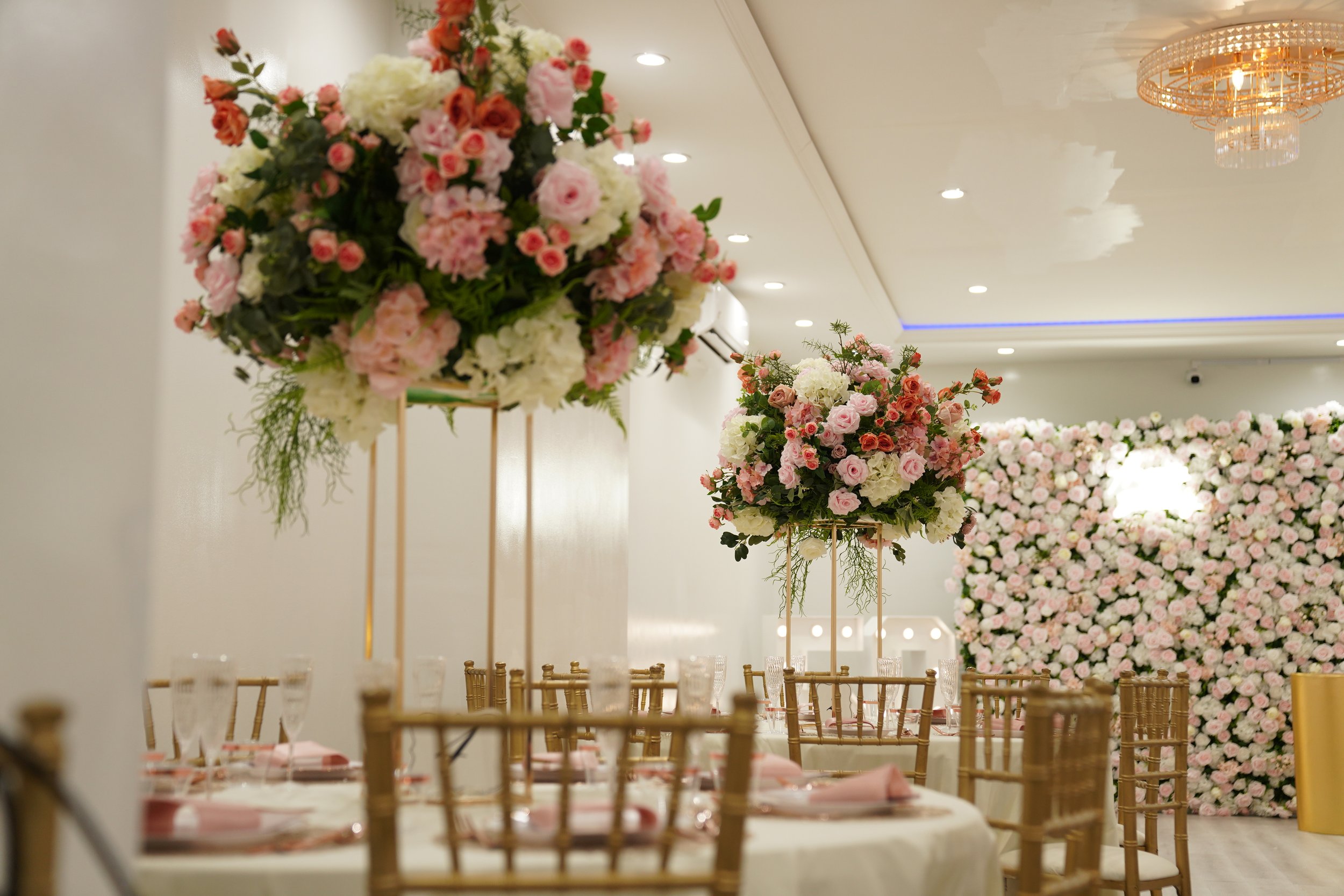 Elegant event space decorated with large floral centerpieces of pink, white, and peach flowers, and a wall of similar pink and white flowers in the background. Gold chairs and tables are set with pink napkins and glassware, under a beautifully lit ceiling with chandeliers.