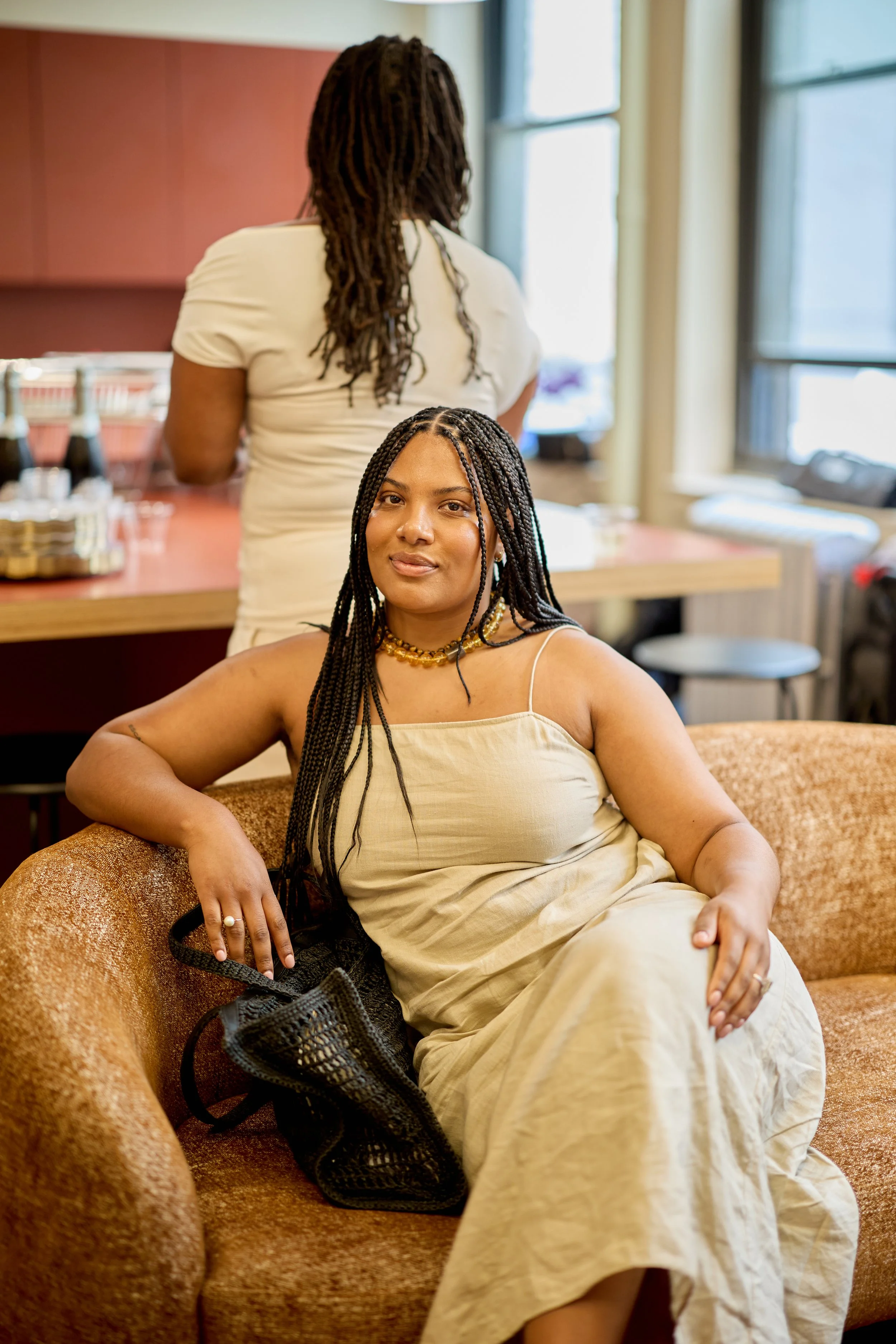 A woman with long braids sitting on a brown sofa in a cozy indoor setting, with another woman with braids standing in the background near a desk with drinks and glasses.