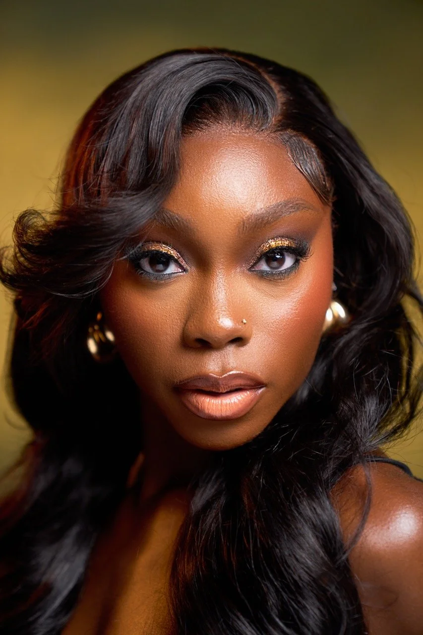 Close-up portrait of a Black woman with styled black hair, gold eyeshadow, and gold earrings, looking directly at the camera.
