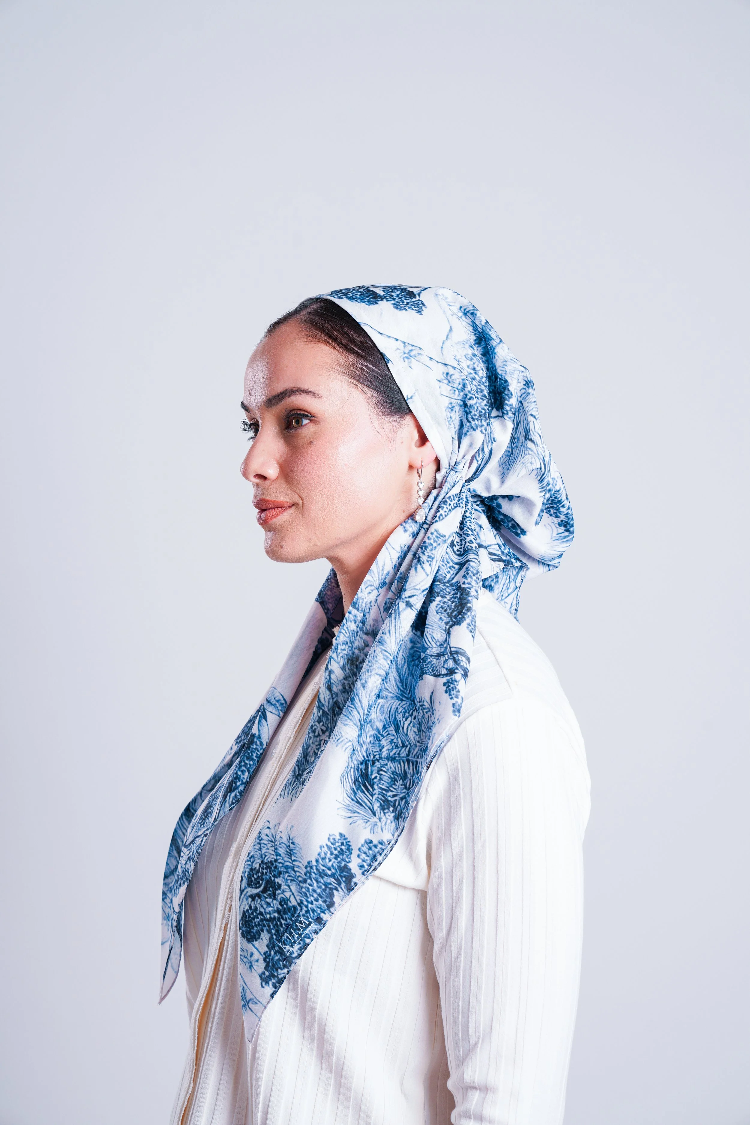 A woman with dark hair wearing a white top and a blue and white patterned headscarf, looking to the left side of the image.