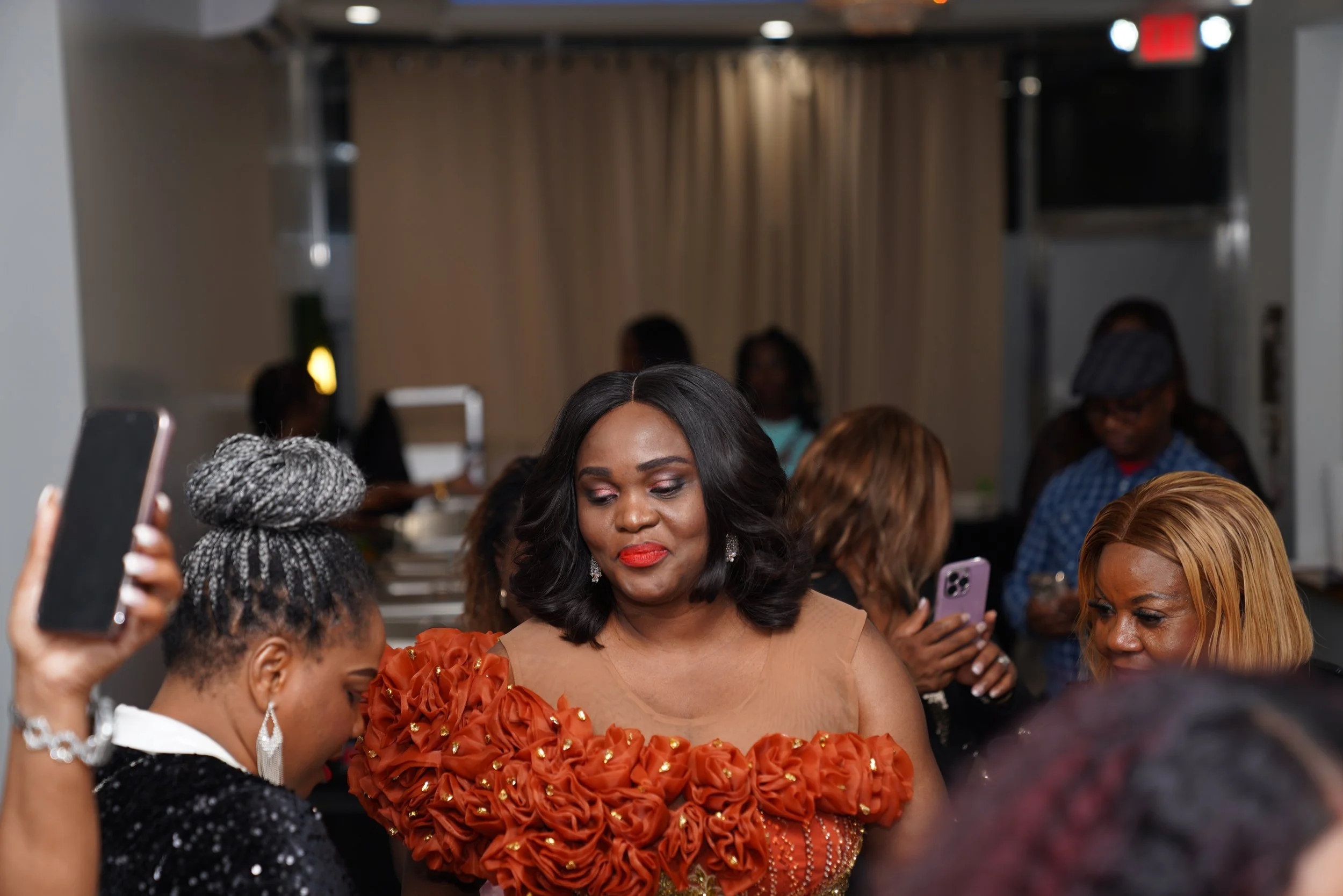 A group of women at a social gathering, some taking photos with their smartphones, with a woman in an ornate orange dress in the center.