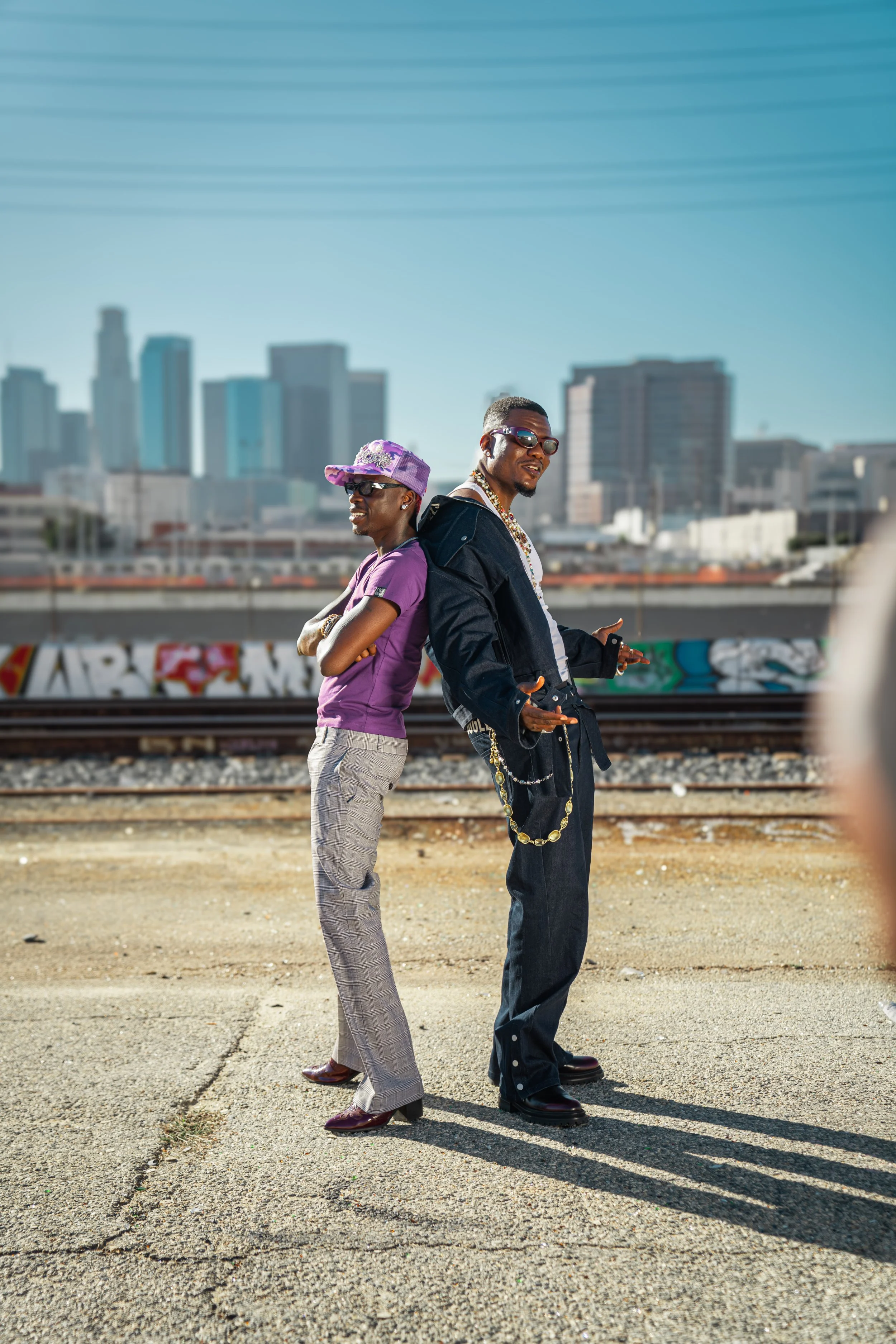 Two men standing back to back near train tracks in an urban area with city skyscrapers in the background, wearing stylish clothing and sunglasses.