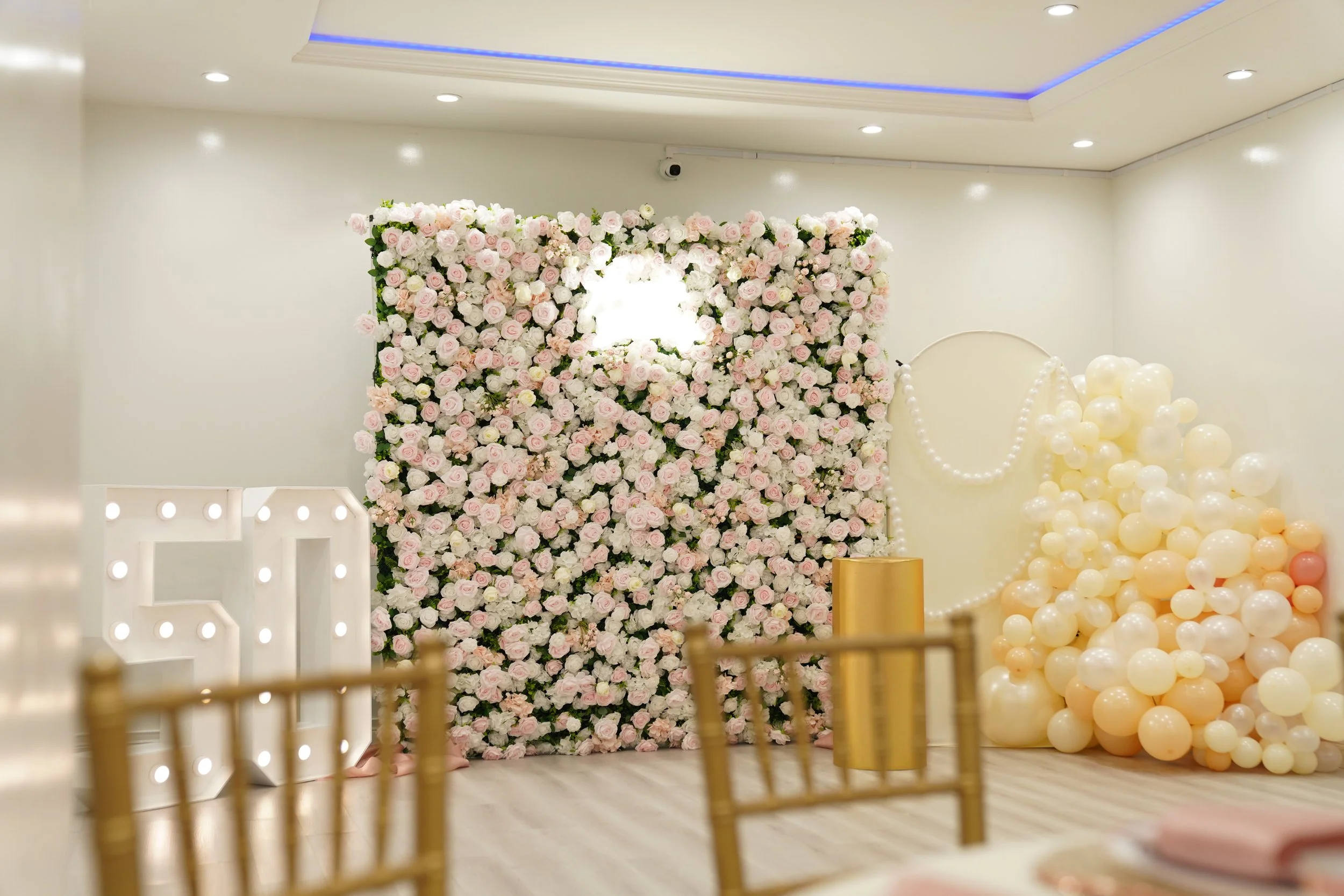 Decorative backdrop with pink and white roses, illuminated letter 'S', and balloons, likely set up for a celebration or event.