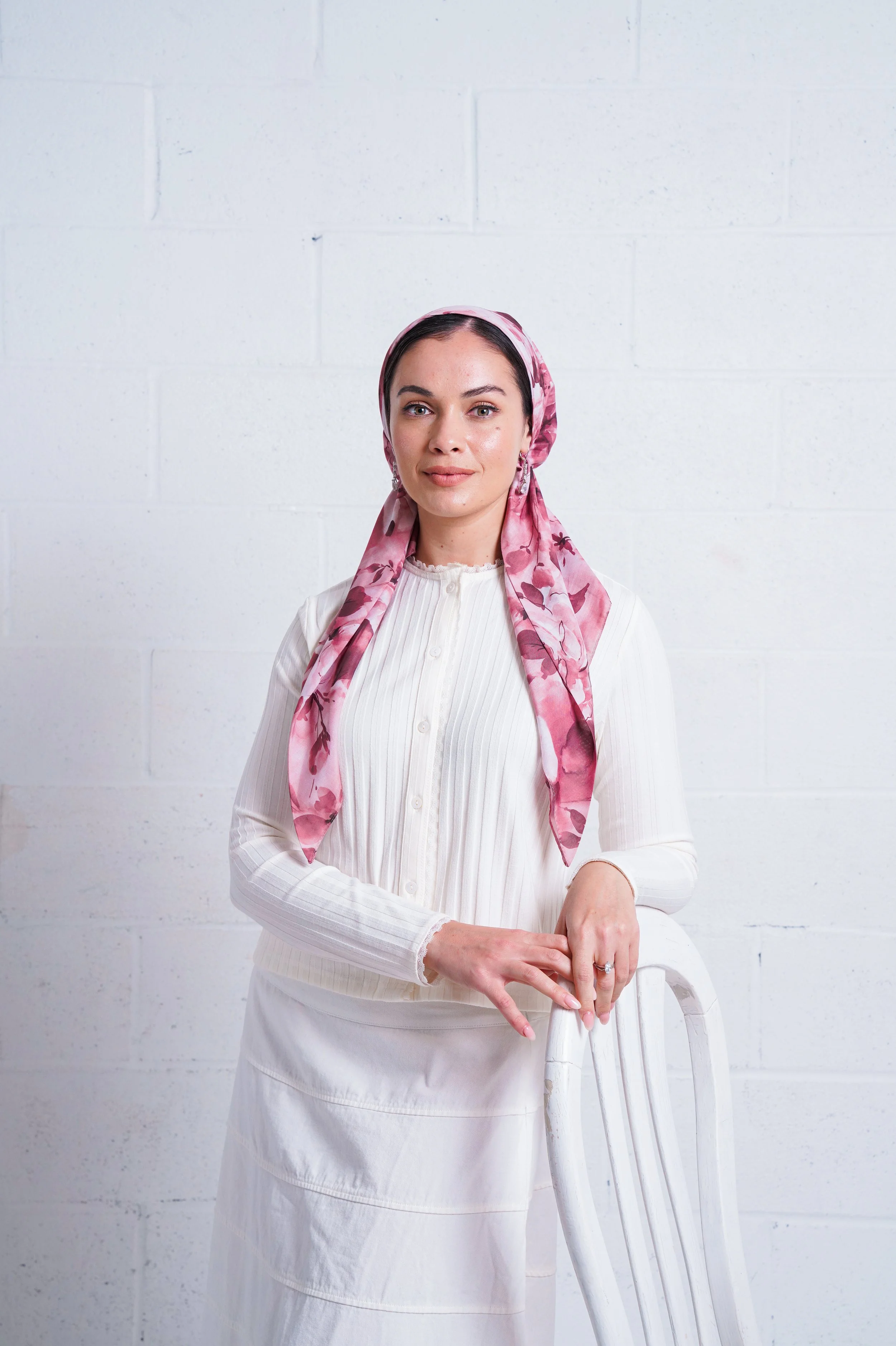Portrait of a woman with fair skin wearing a light pink floral headscarf, standing against a white brick wall, dressed in a white button-up top and skirt, with her hand resting on a white wooden chair.