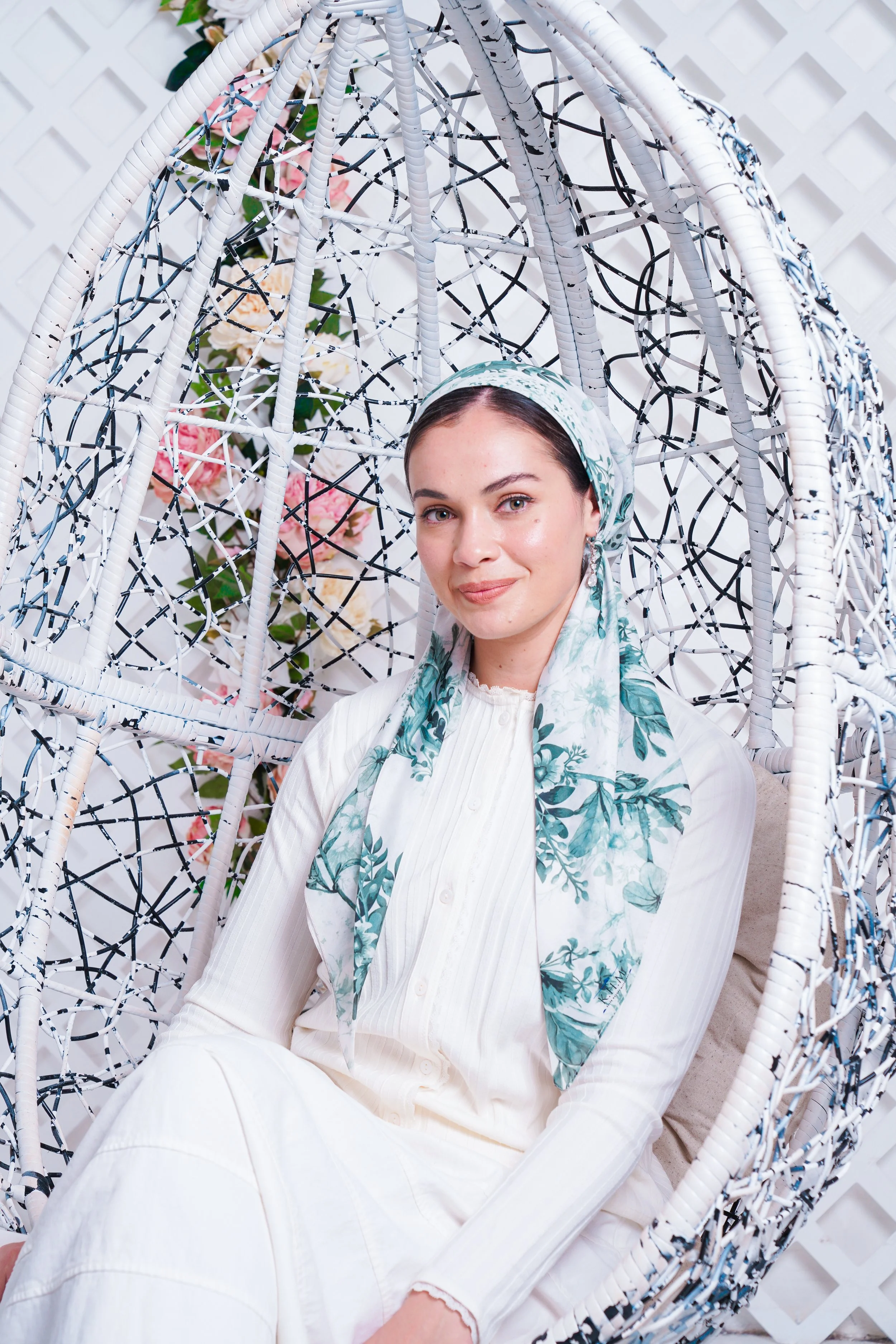 A woman sitting in a hanging chair with a floral backdrop, wearing a white outfit and a green patterned headscarf.