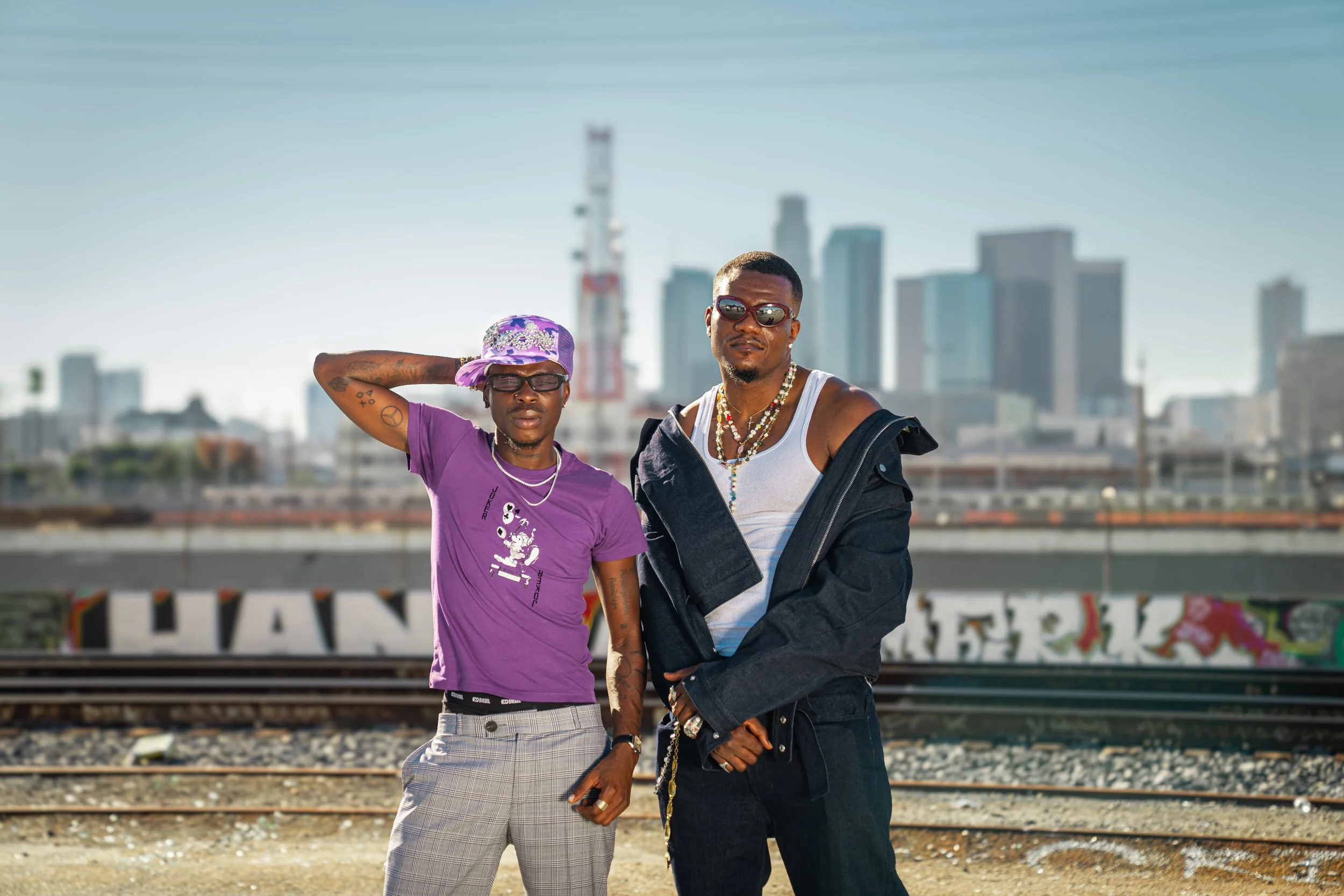 Two young men standing on a railway track with a city skyline in the background. One is wearing a purple T-shirt, plaid pants, a purple hat, sunglasses, and has tattoos. The other is wearing a white tank top, black jacket, multiple necklaces, and sun