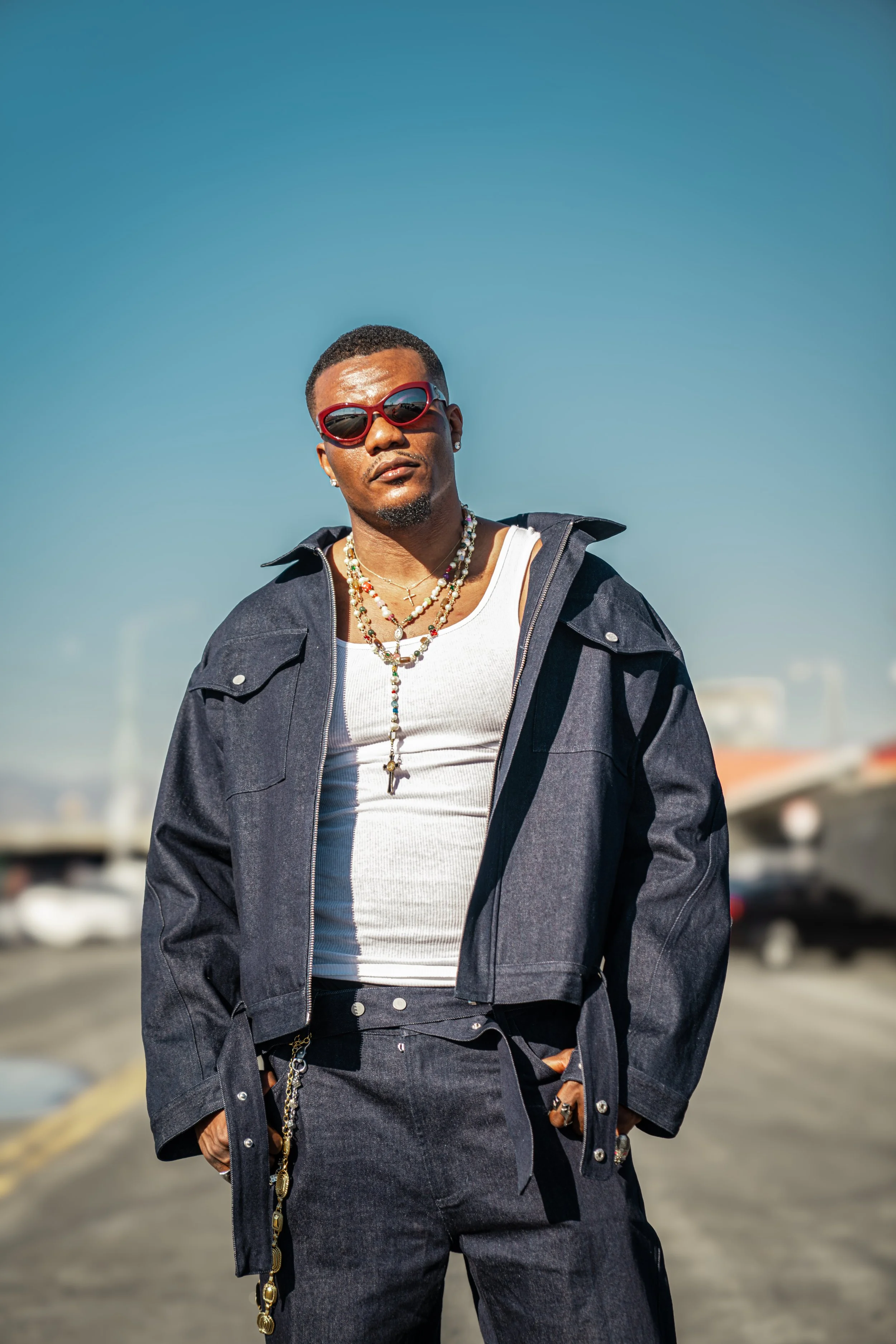 A man wearing a dark denim jacket over a white tank top, with colorful beaded necklaces and sunglasses, standing outdoors on a sunny day with boats and sky blurred in the background.