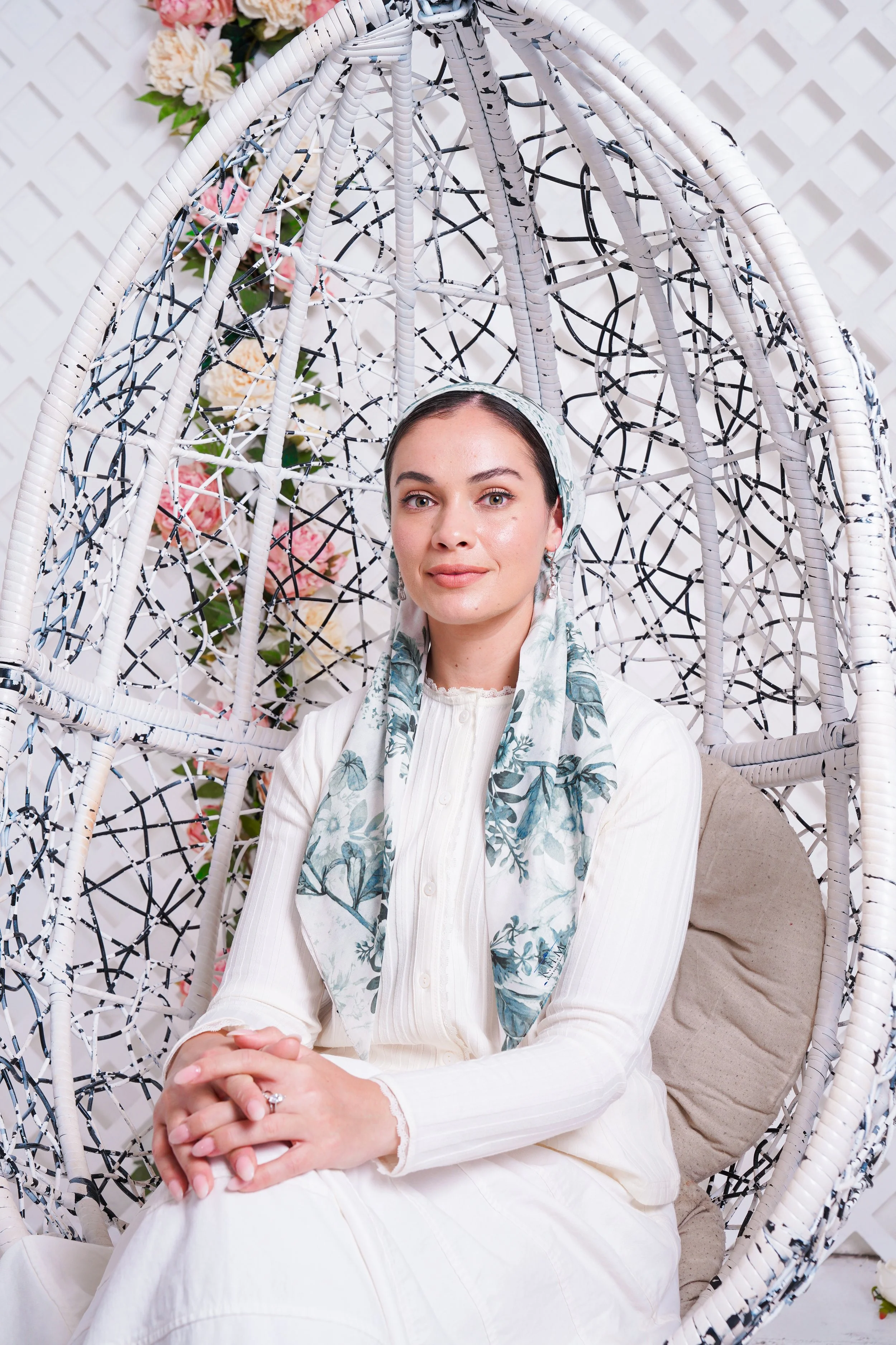 Woman sitting in a white woven hanging chair with a floral scarf, white blouse, and a beige cushion, in front of a floral background.
