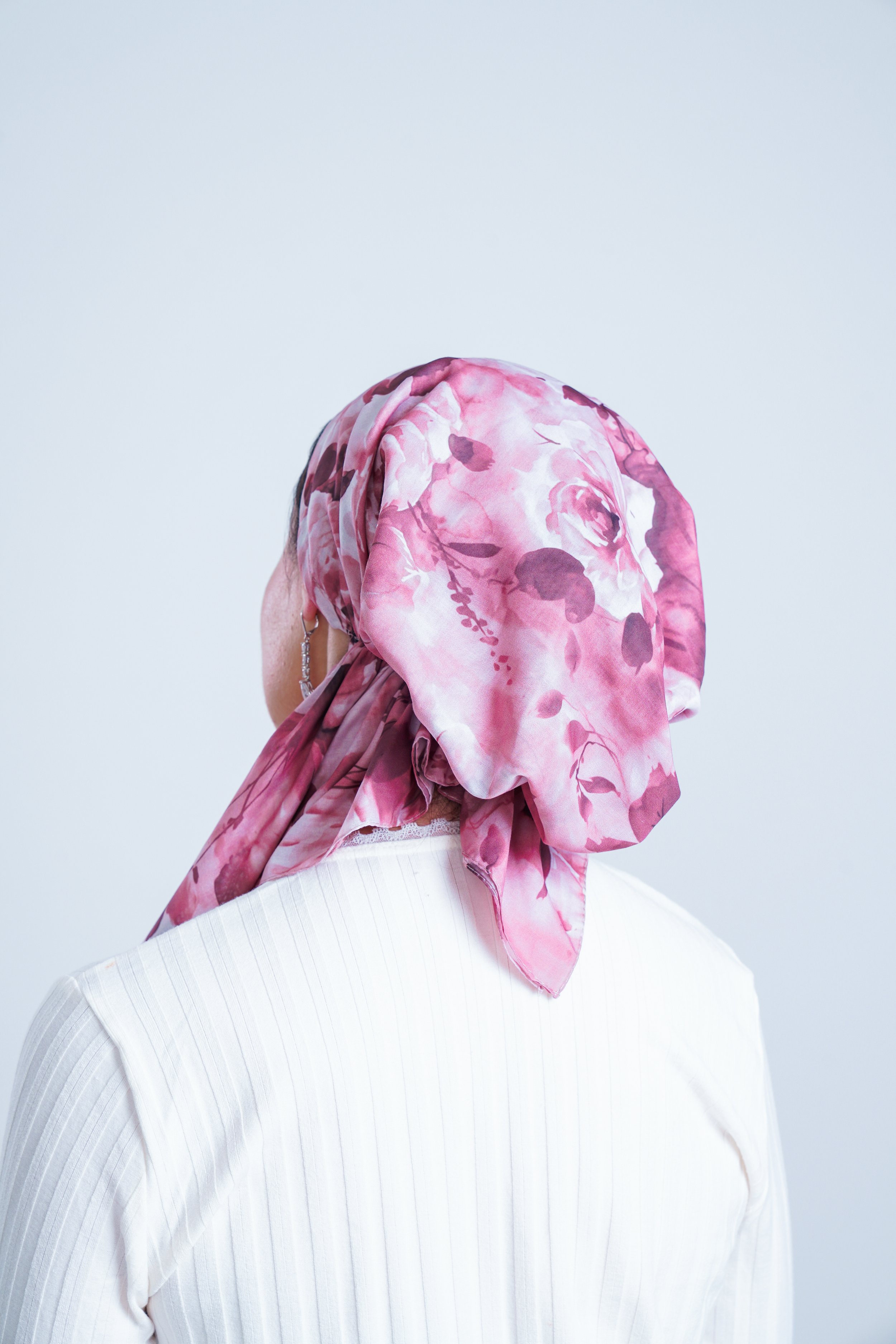 A person wearing a pink floral headscarf and a white shirt, facing sideways against a plain light background.
