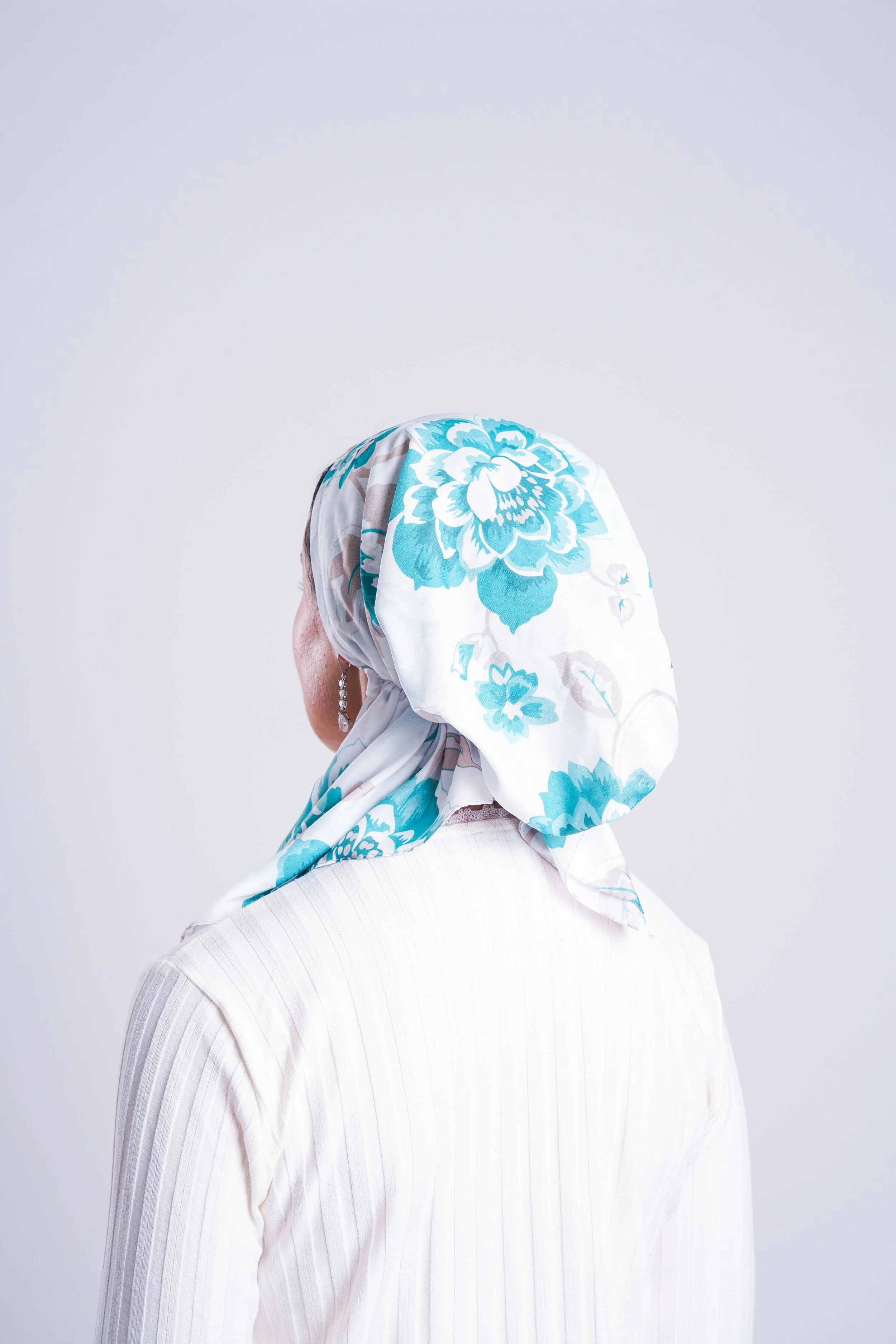 A woman wearing a white blouse with subtle stripes and a floral headscarf with large light blue flowers on a white background, seen from the side.