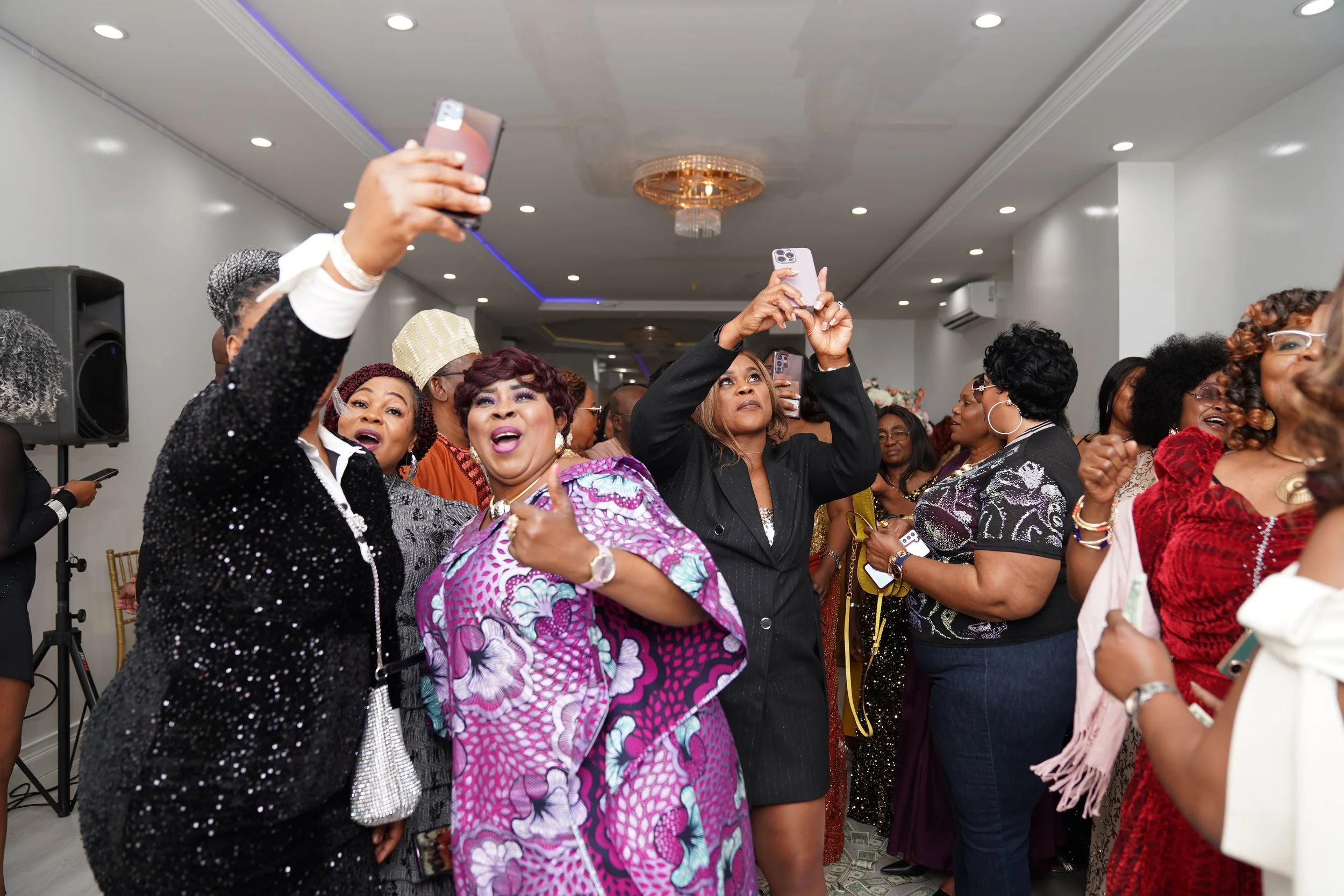 Group of women at a celebration or party, taking photos with smartphones, dressed in colorful and elegant attire.
