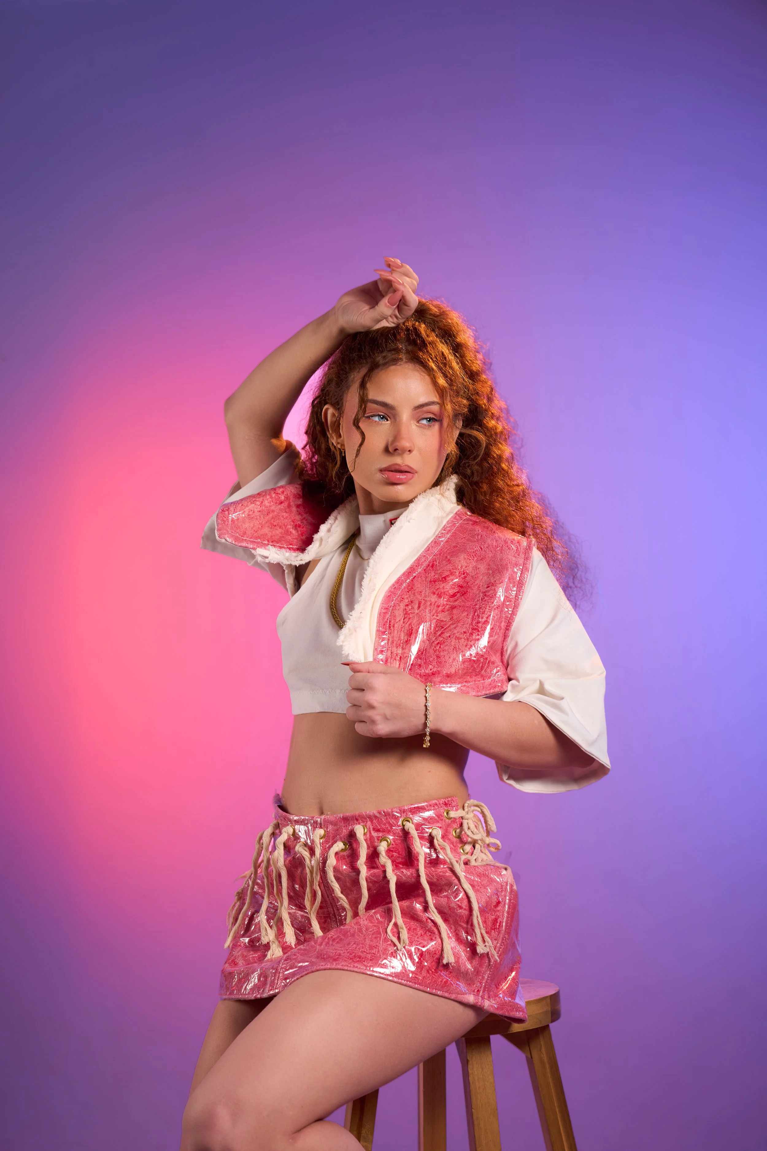 A young woman with curly red hair posing against a purple and pink gradient background. She is sitting on a wooden stool, wearing a pink shiny mini skirt with tassels, a white cropped top, and a pink vest. She has one arm raised above her head and the other holding the vest, with a focused expression on her face.