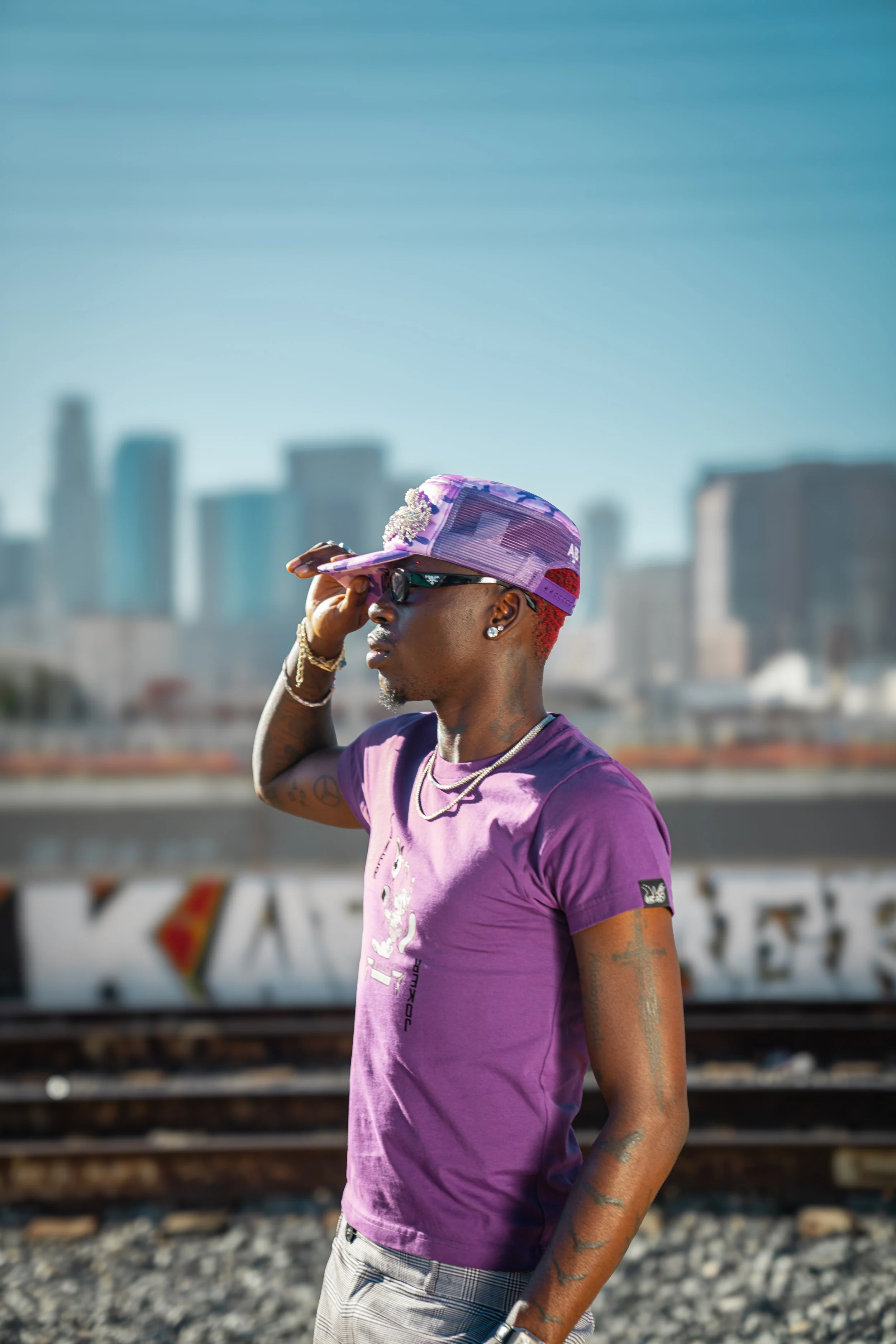 A man wearing a purple t-shirt, purple cap, and sunglasses standing outdoors near train tracks with a city skyline in the background.
