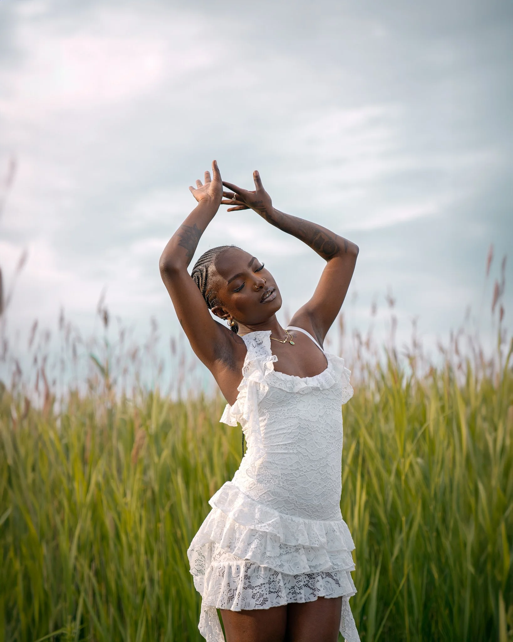 A woman with braided hair and tattoos on her arms is standing in a field of tall grass, wearing a white lace dress, with her arms raised above her head, eyes closed, in a peaceful pose.