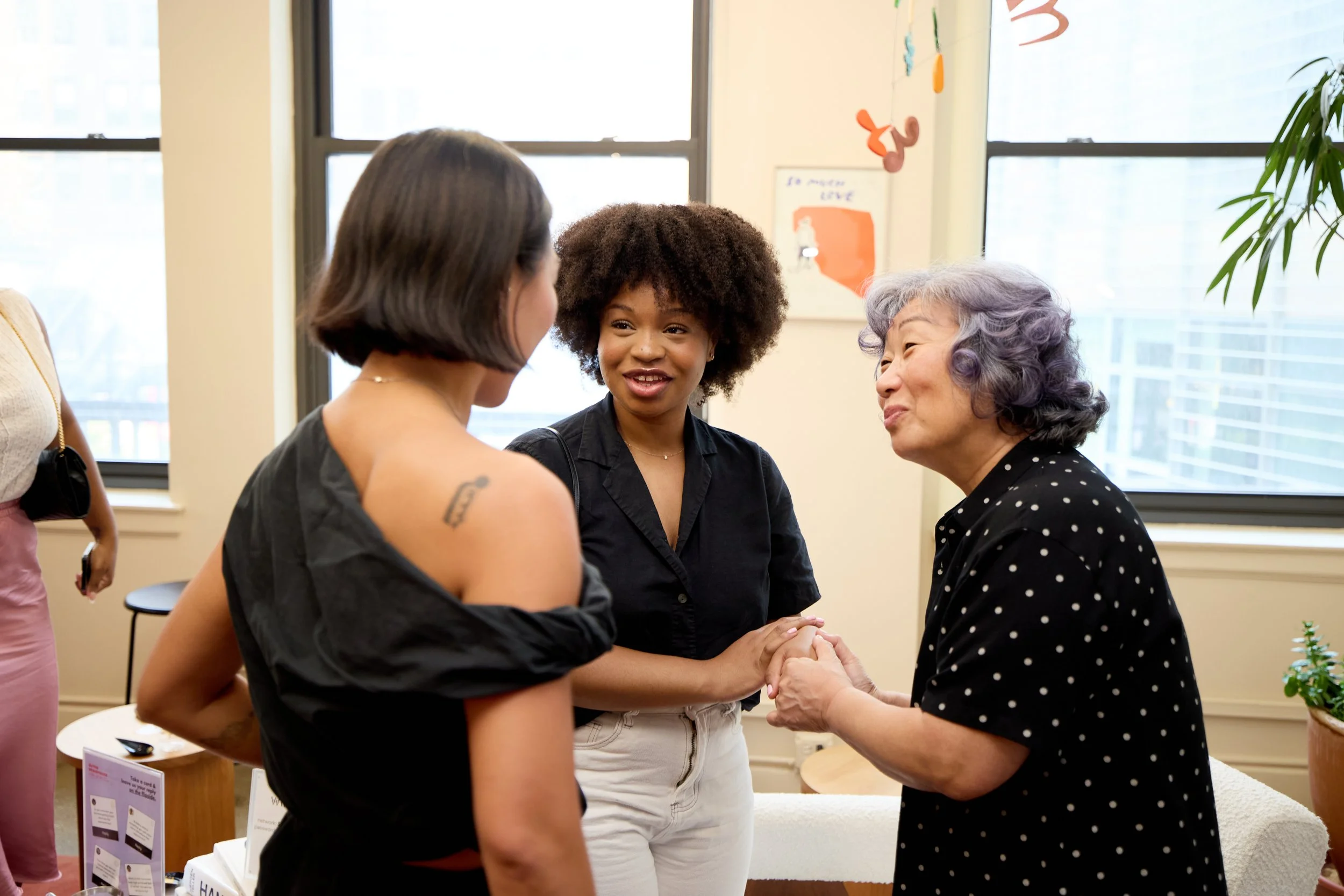 Three women talking and holding hands in a bright room with windows, plants, and wall decorations.