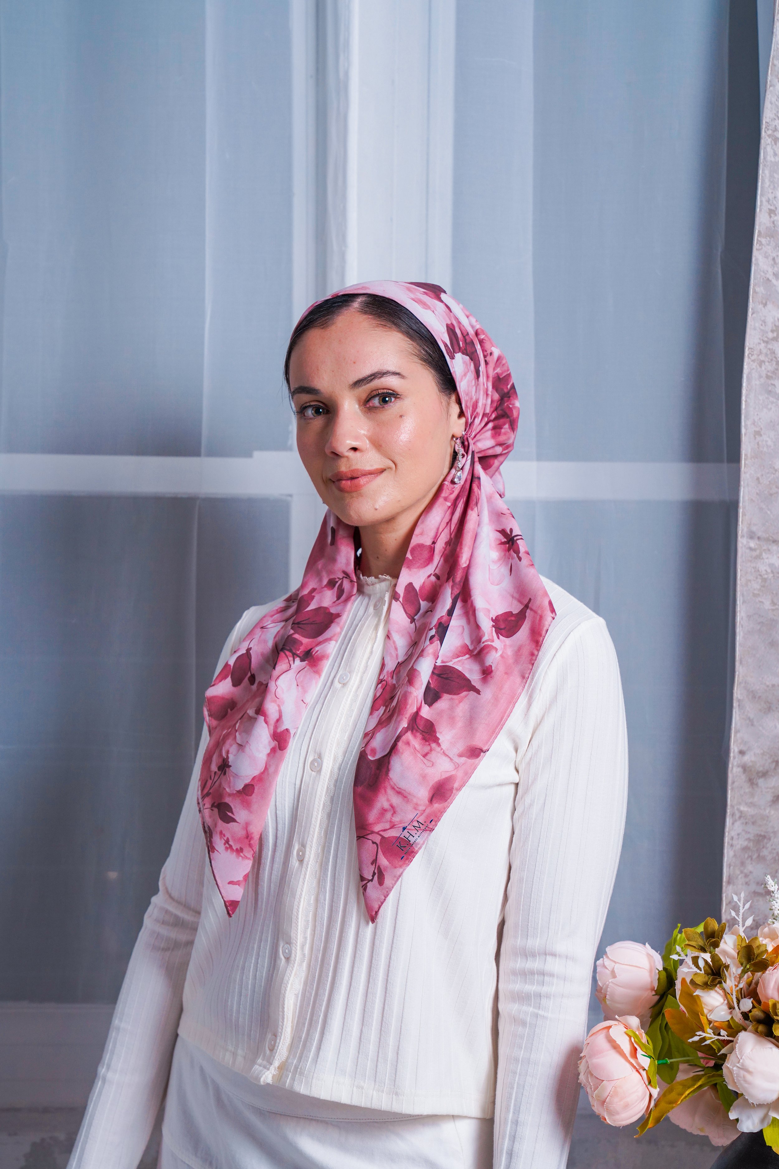 A young woman with fair skin and dark hair, wearing a pink floral headscarf and a white ribbed button-up top, standing in front of a light background with floral accents.