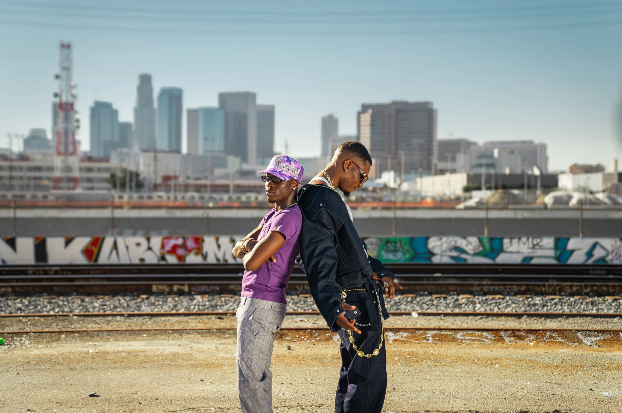 Two young men stand back to back on a graffiti-covered area near train tracks in an urban setting, with a city skyline in the background during daylight.