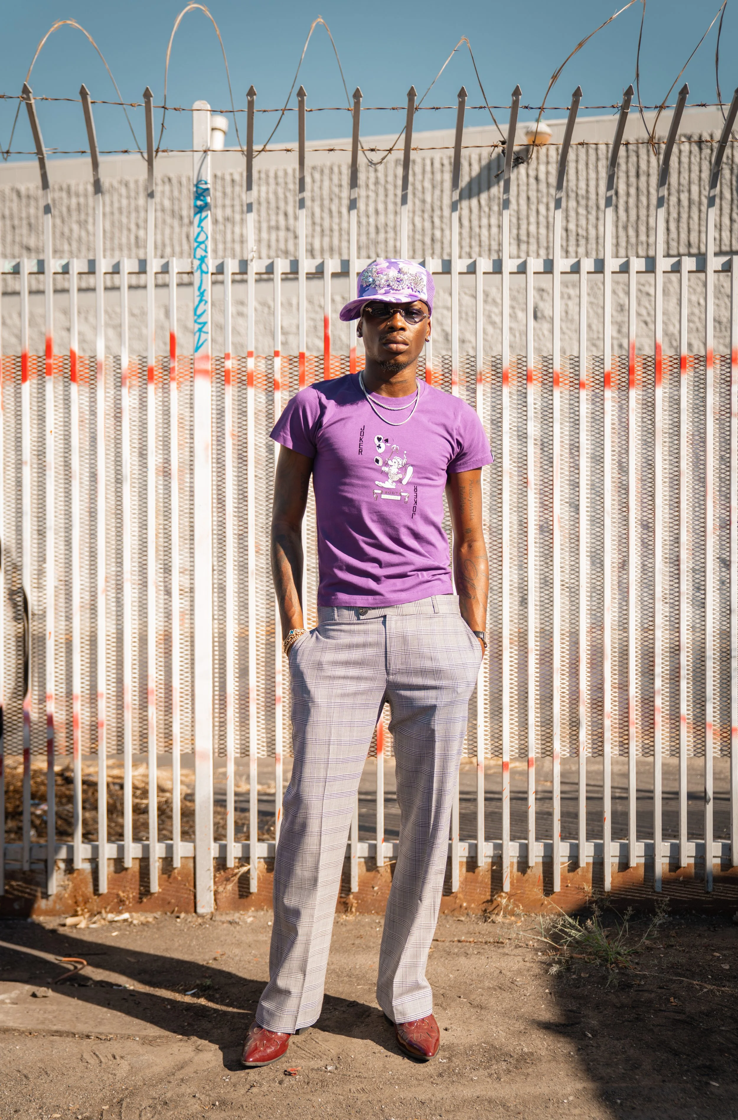 A man in a purple t-shirt, checkered gray pants, a purple cap, and sunglasses standing in front of a metal fence with barbed wire on top, outdoors on a sunny day.