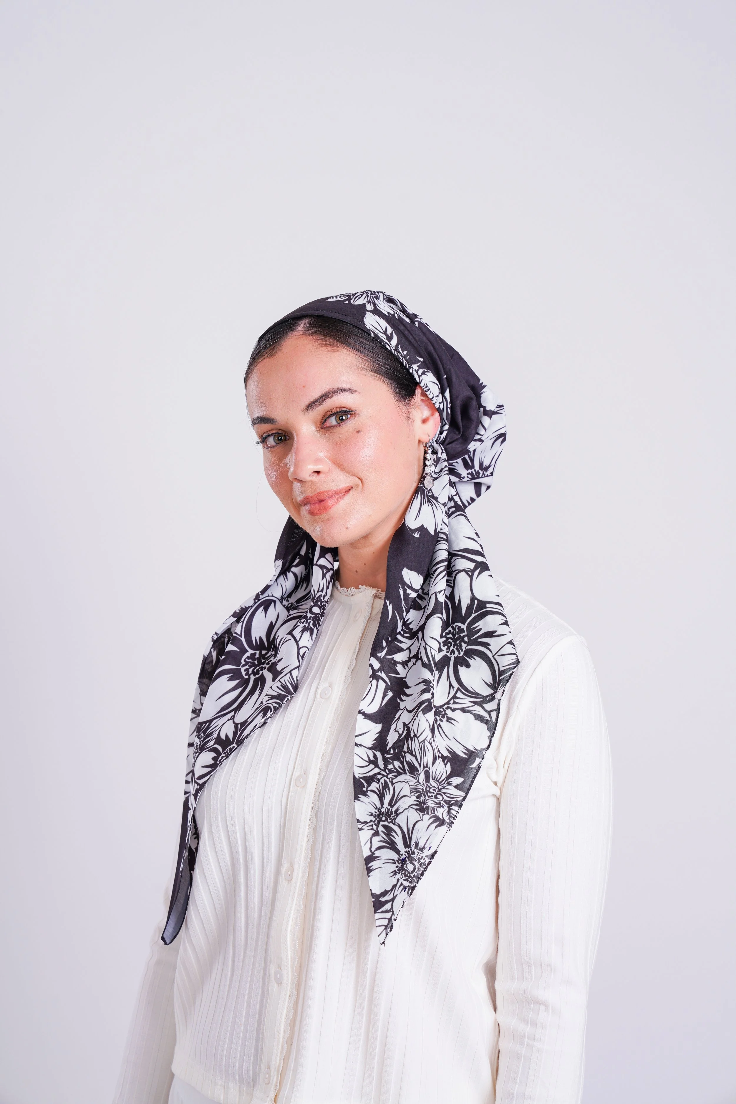 A woman wearing a black and white floral headscarf and a white button-up sweater, smiling at the camera against a plain light background.