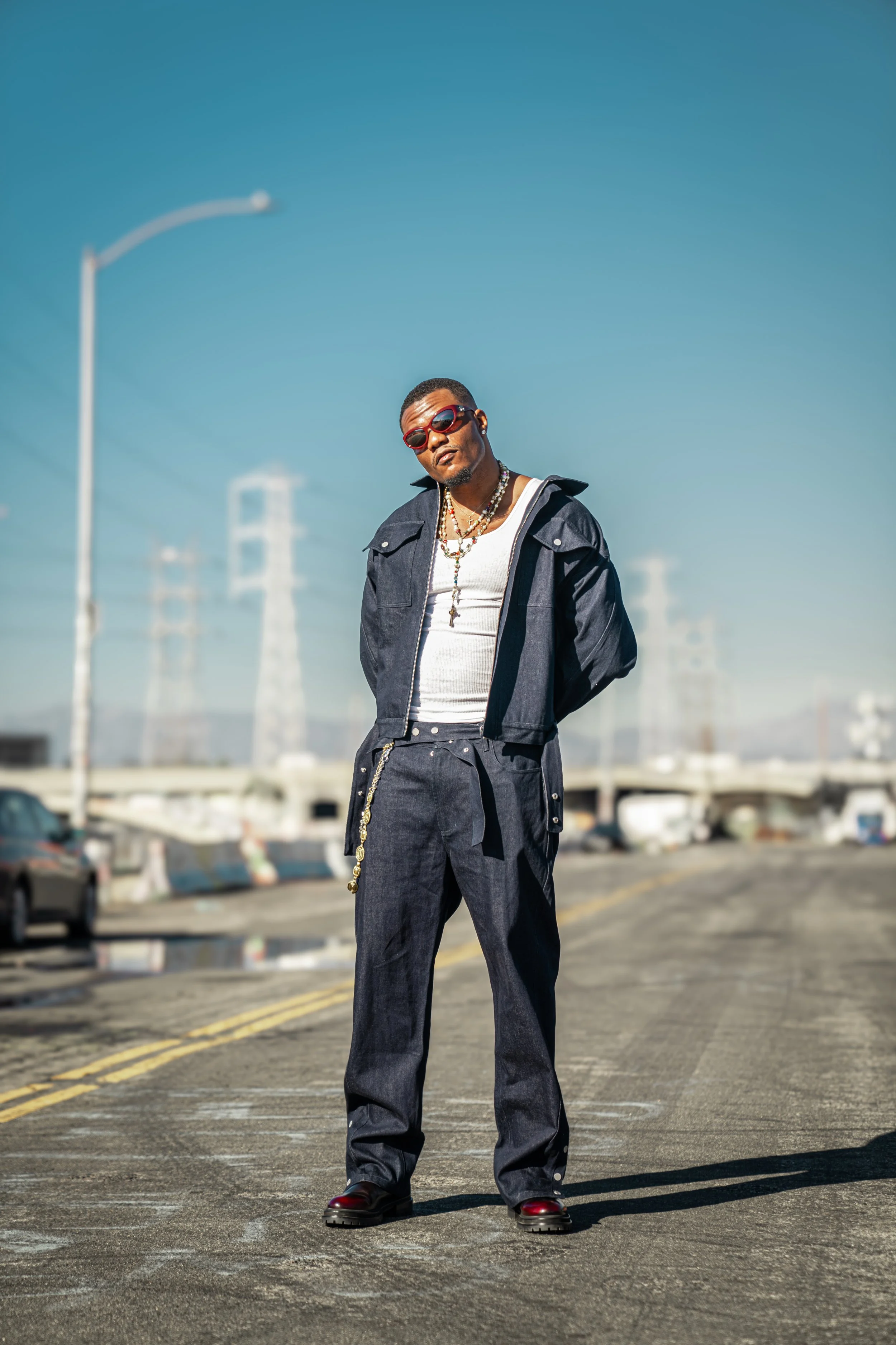 A man wearing sunglasses, a white tank top, a dark jacket, matching baggy jeans with a chain, and boots standing on a street with power lines in the background and a clear sky.