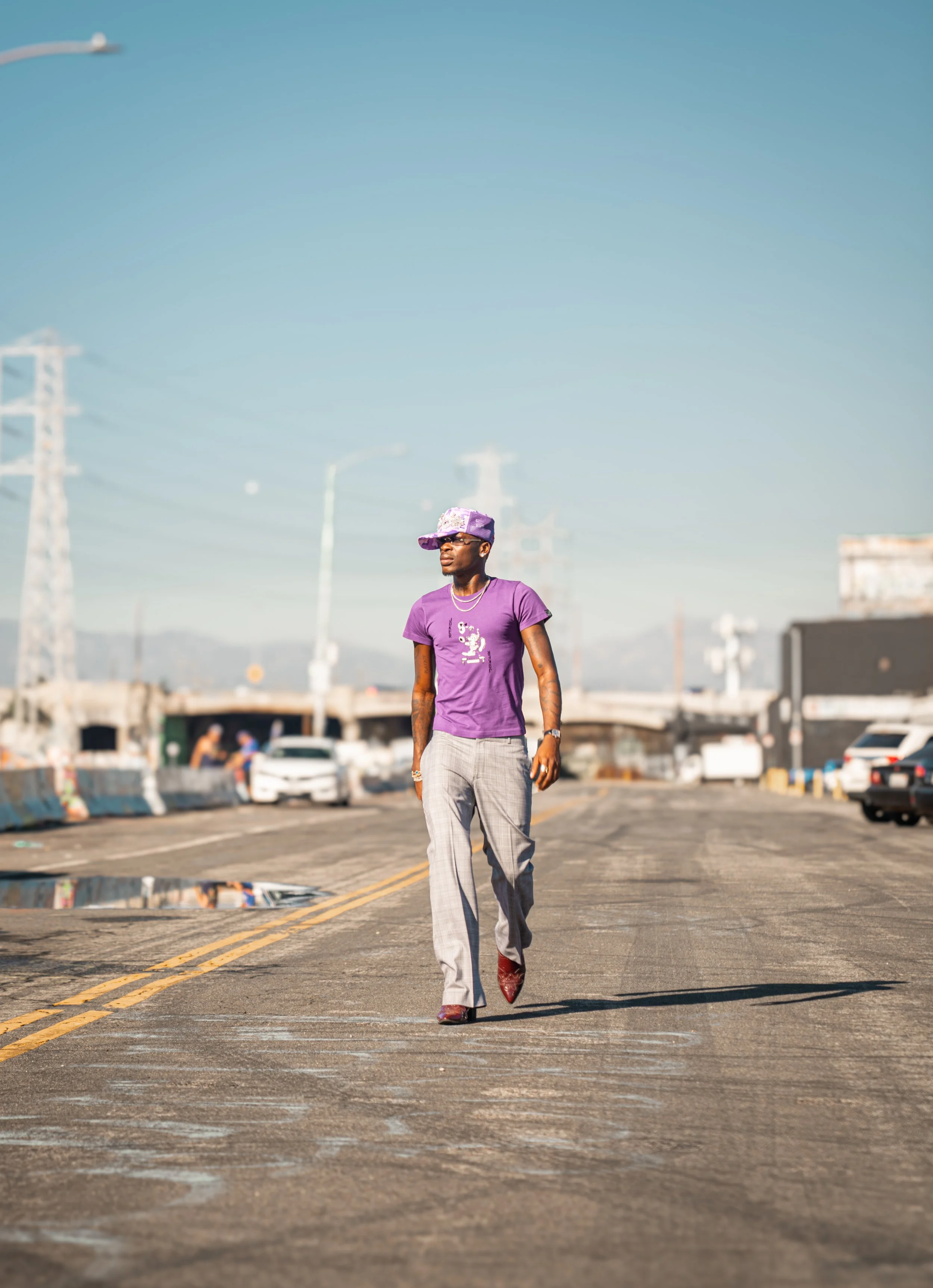 A man walking on a city street wearing a purple t-shirt, plaid pants, and purple hat, with cars and industrial infrastructure in the background on a sunny day.