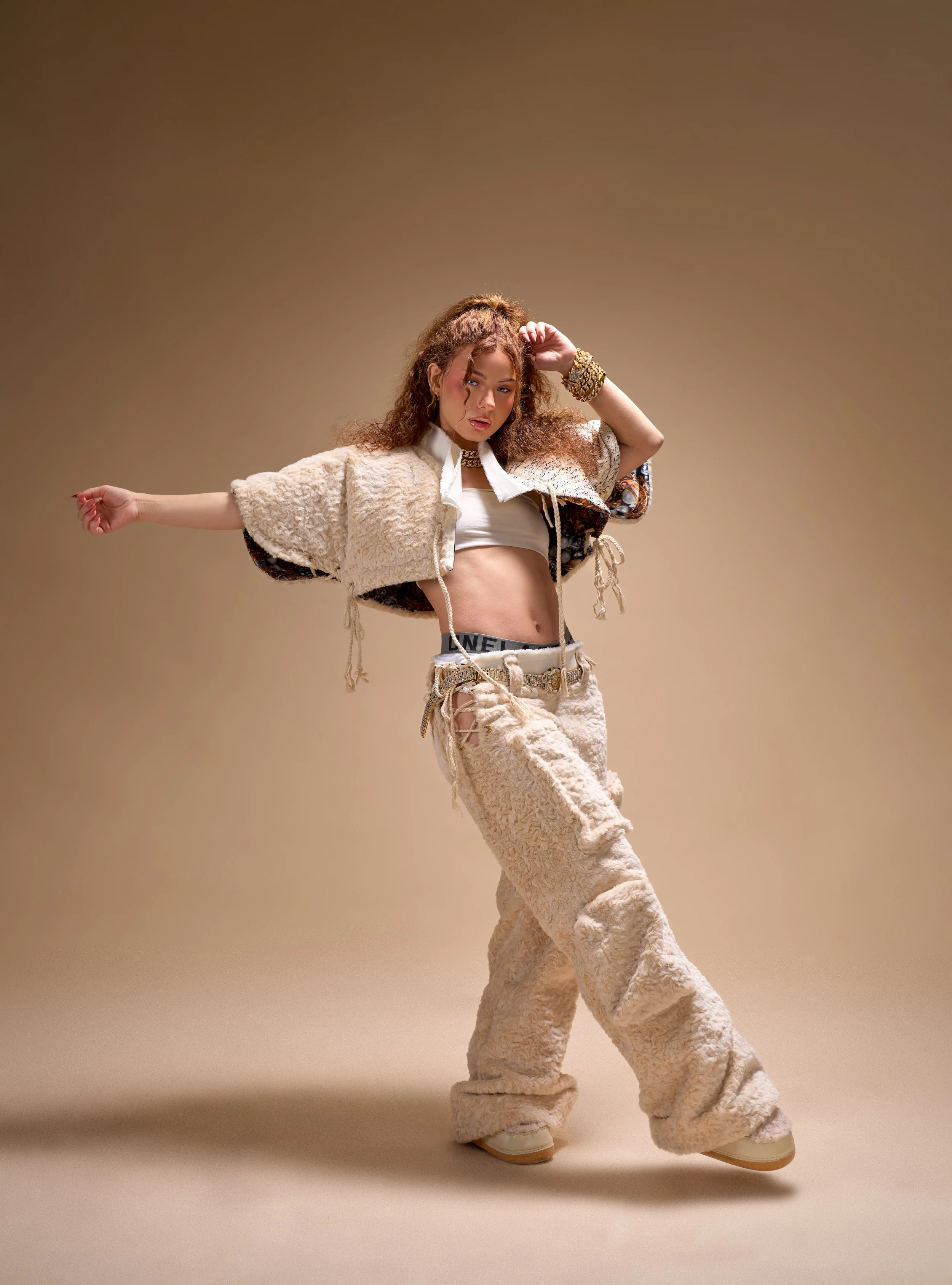 A woman with curly hair posing in a studio, wearing a beige fuzzy jacket and pants, white crop top, and gold accessories.