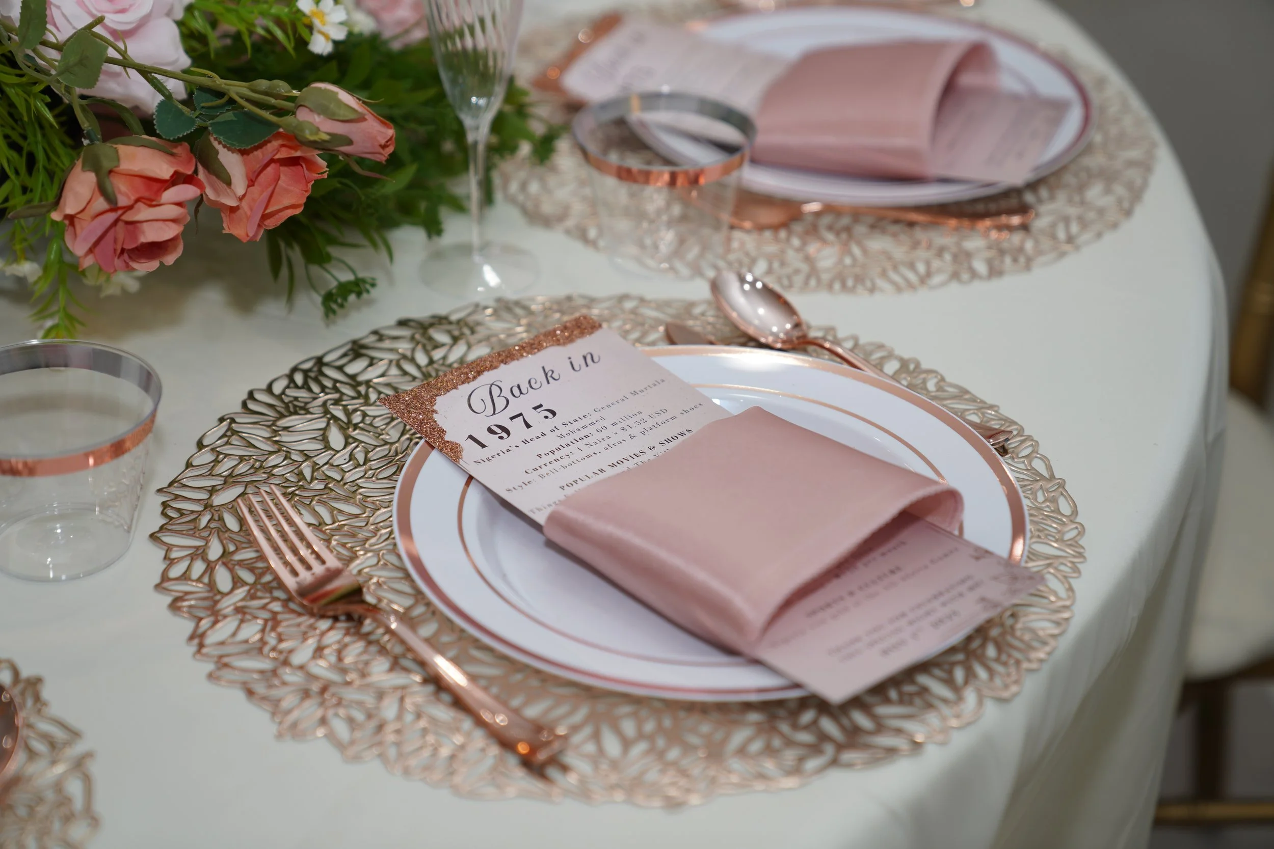 Elegant table setting with pink and gold accents, including a pink napkin, a menu, rose gold cutlery, a clear glass, a floral centerpiece with pink roses, and a gold decorative placemat.