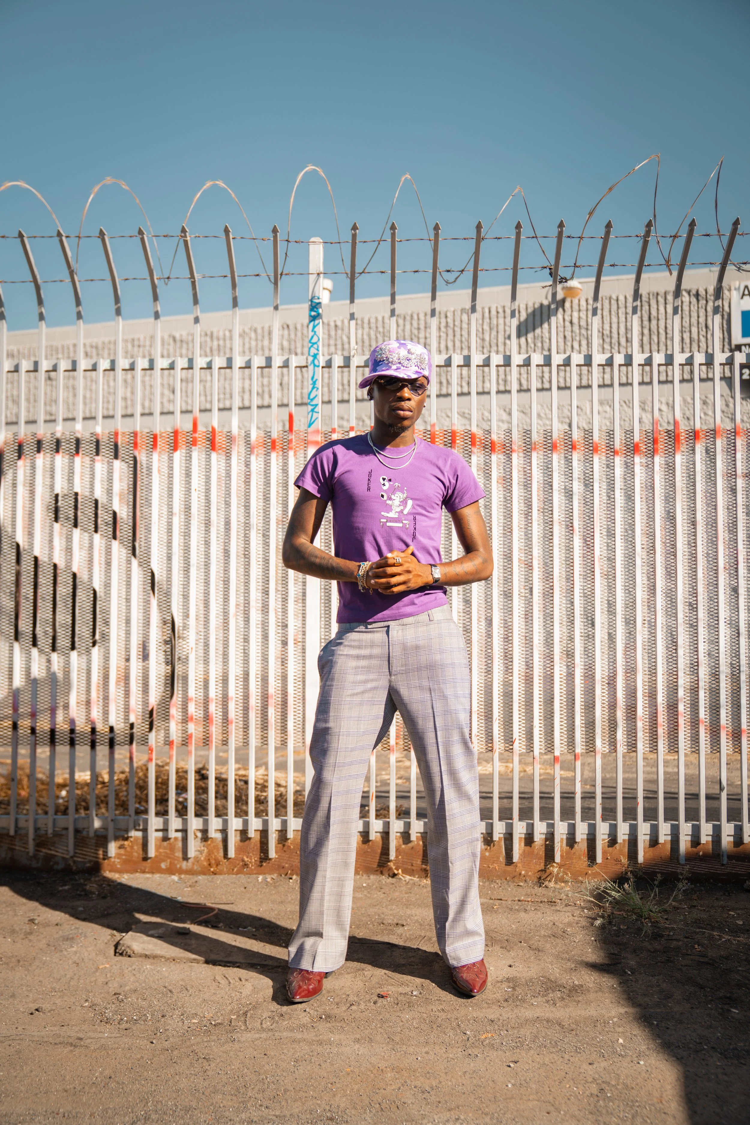 A person standing in front of a barbed wire fence wearing a purple t-shirt, plaid gray pants, red shoes, a hat, sunglasses, and jewelry.