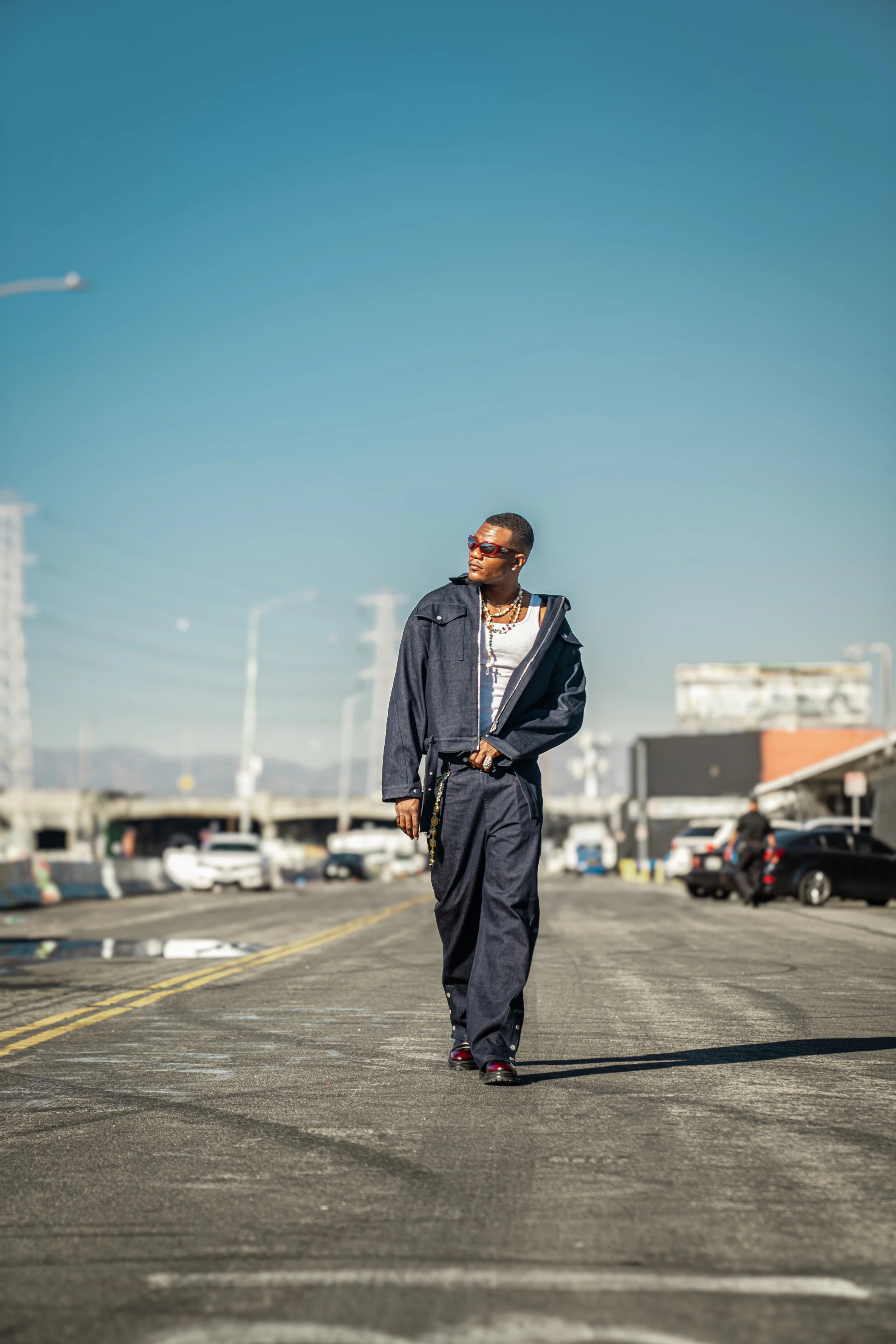 A person walking on an empty parking lot under a clear blue sky, wearing sunglasses, a dark jacket, and loose pants.