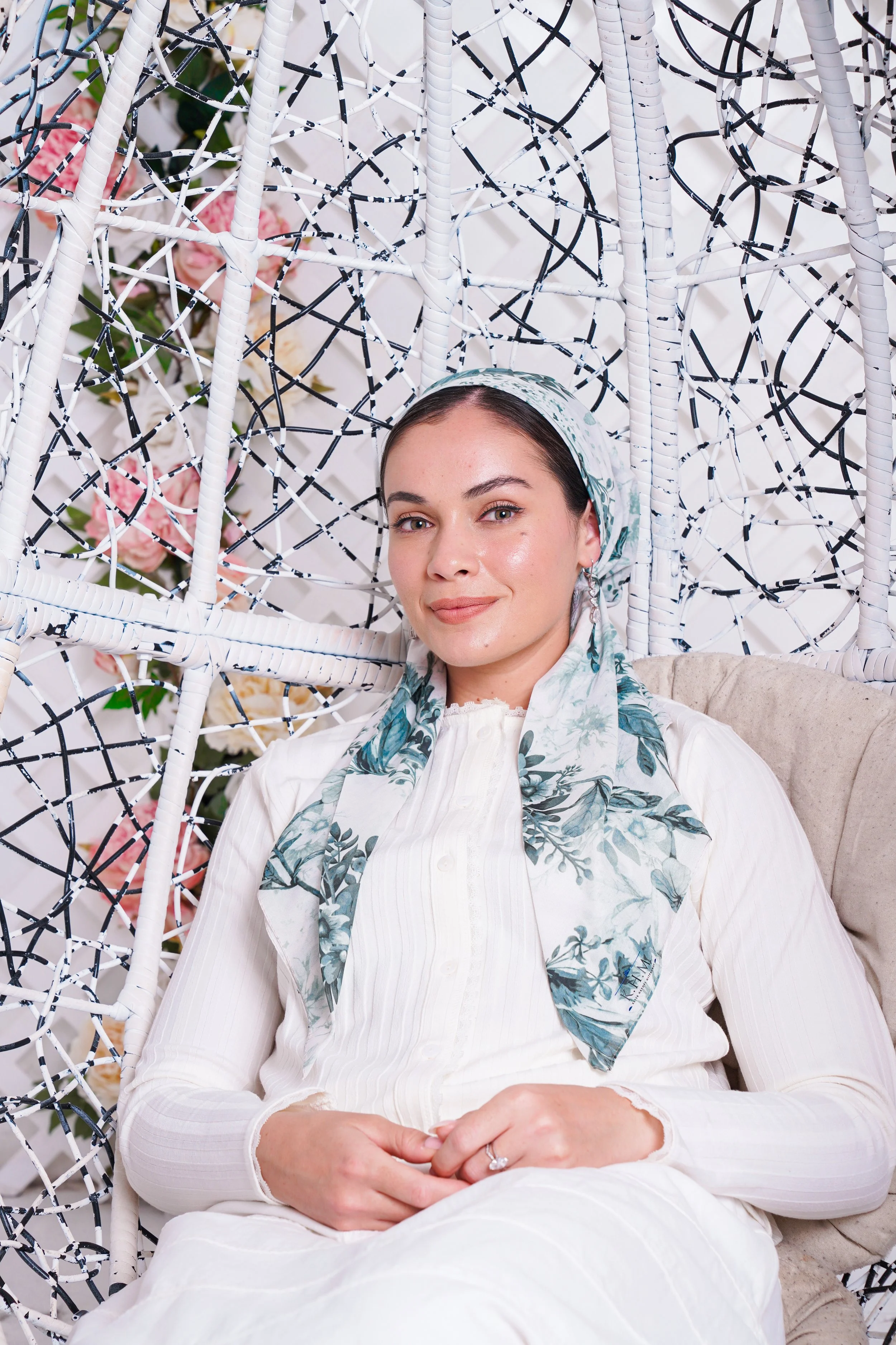 A woman with light skin and dark hair, wearing a floral headscarf and white clothing, sitting on a beige chair in front of a decorative white and black lattice structure with pink roses in the background.