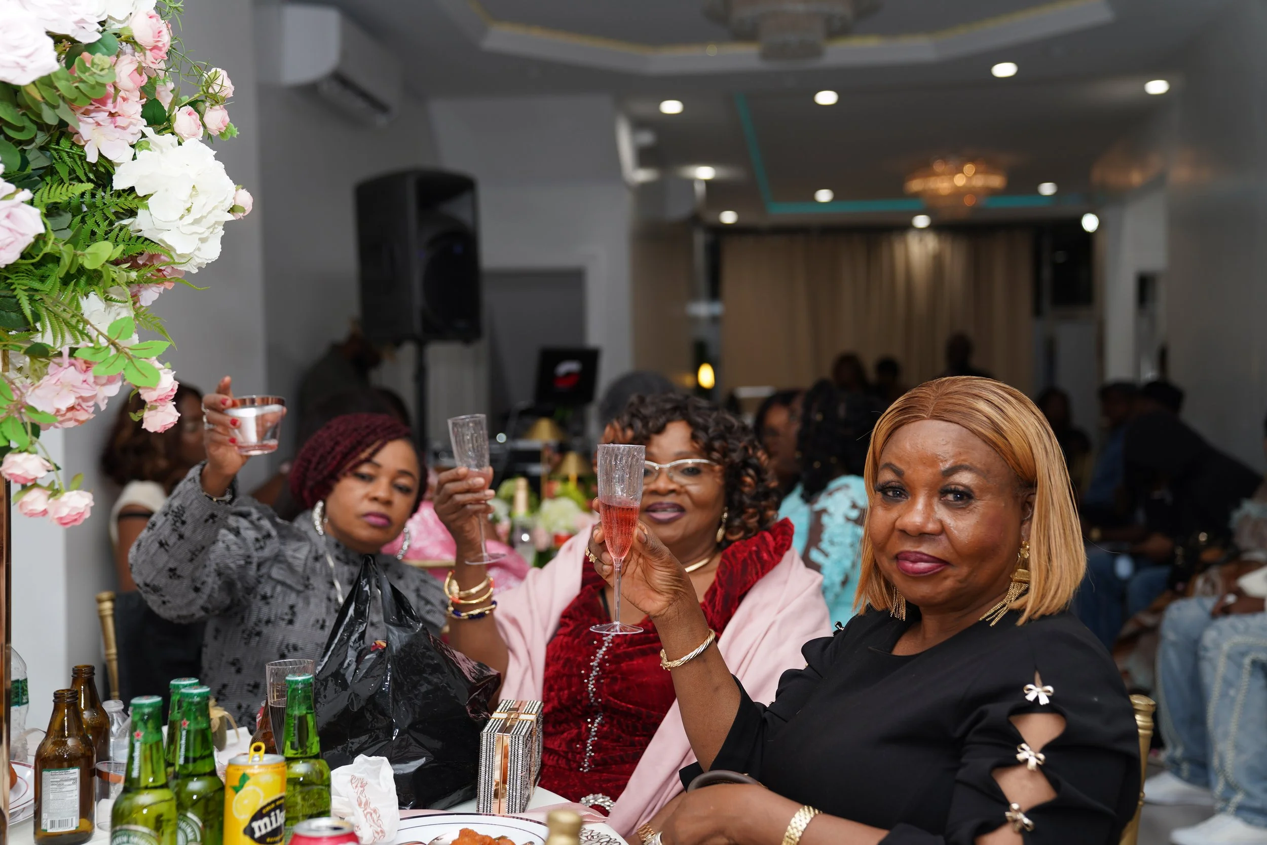 Women at a celebration raising glasses at a decorated banquet table, with bottles and food, in a banquet hall.