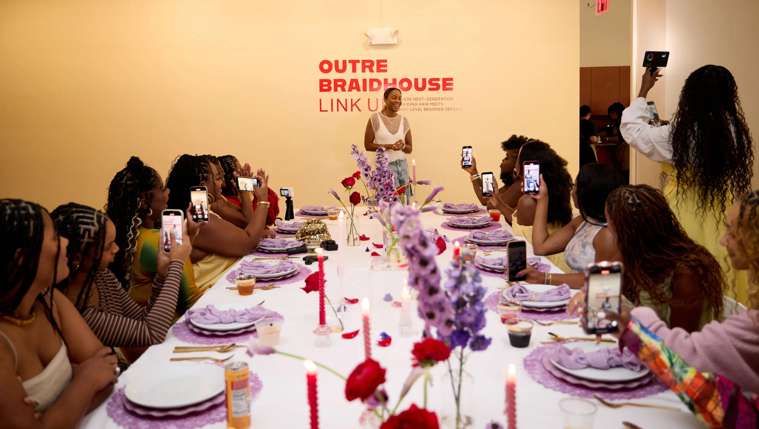 A woman standing and smiling in front of a table set for a gathering with women taking photos and videos of her. The background shows a sign that reads "OUTRE BRAIDHOUSE LINK UP".