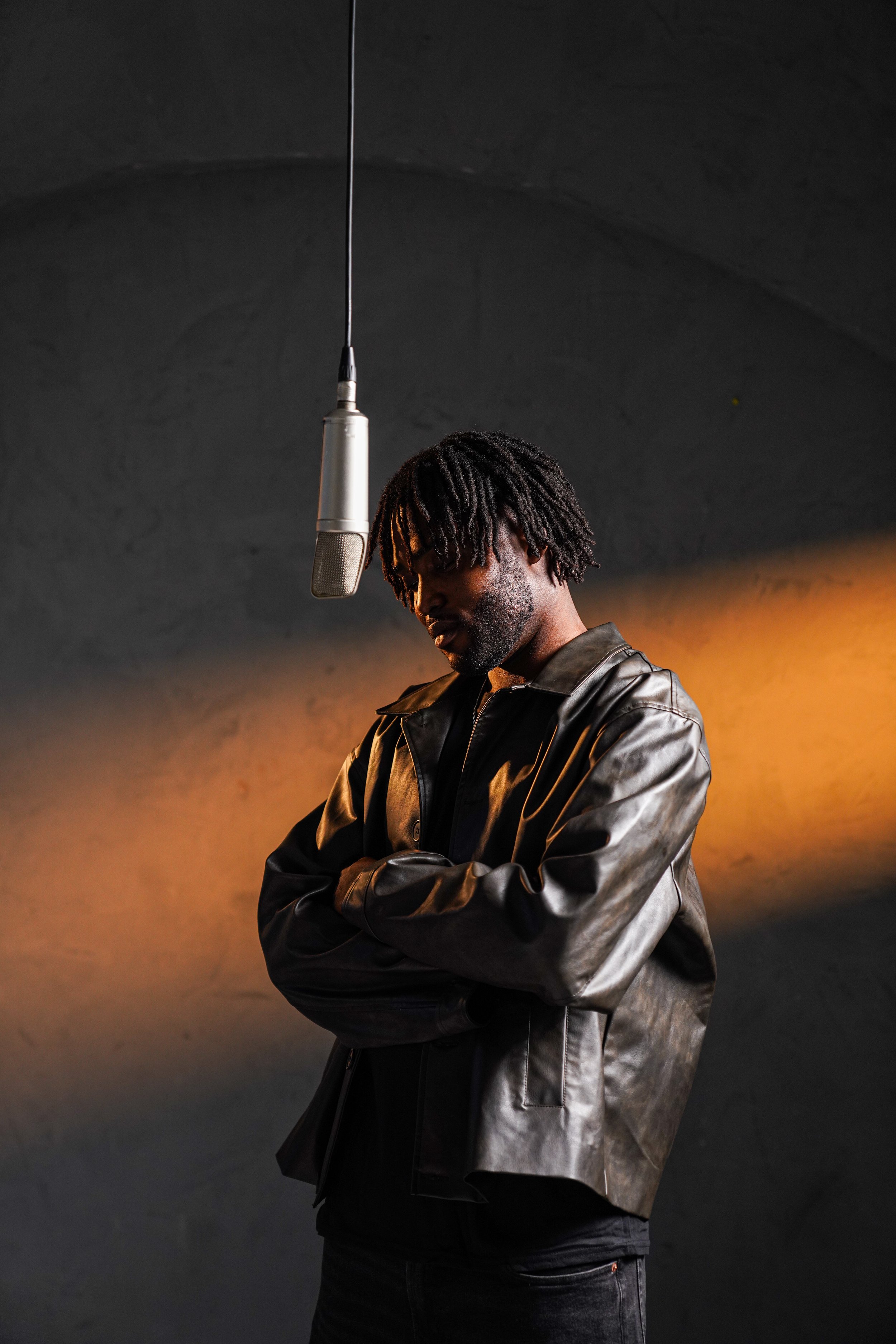 A man in a black leather jacket standing with arms crossed, looking down, with a microphone hanging from the ceiling overhead, in a dimly lit room with a warm orange glow on the wall behind him.