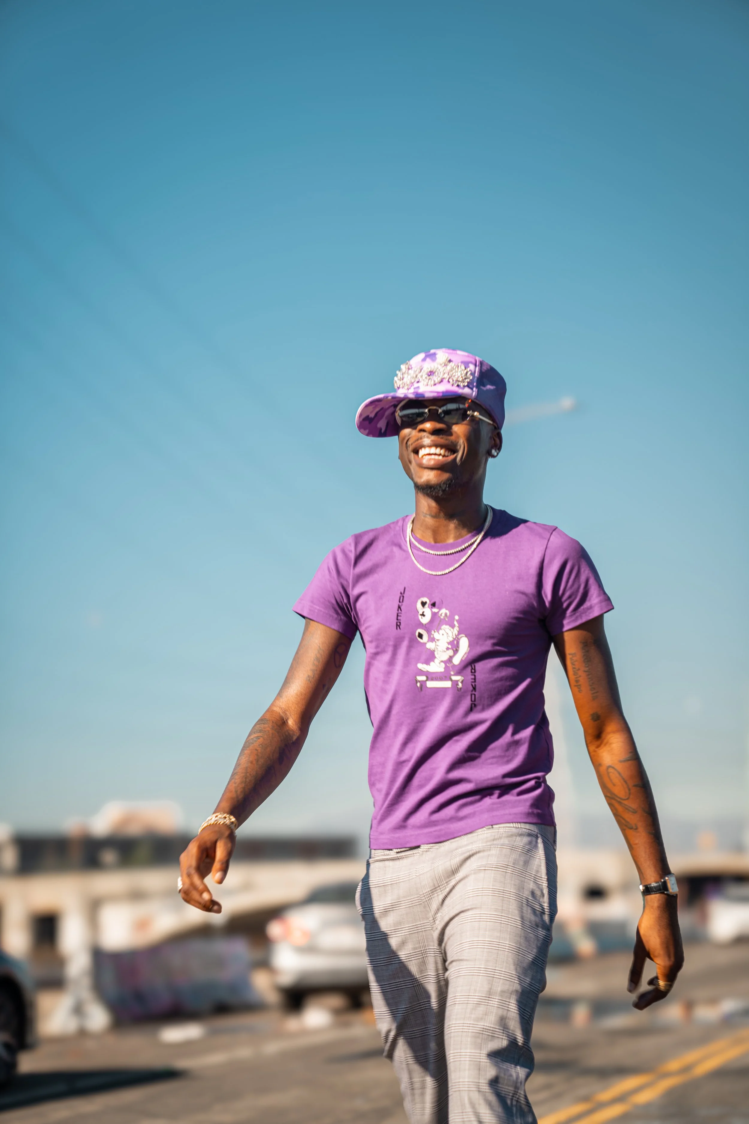 A man smiling while walking outdoors on a sunny day, wearing a purple T-shirt with a graphic design, plaid pants, a purple cap with embellishments, sunglasses, and jewelry.