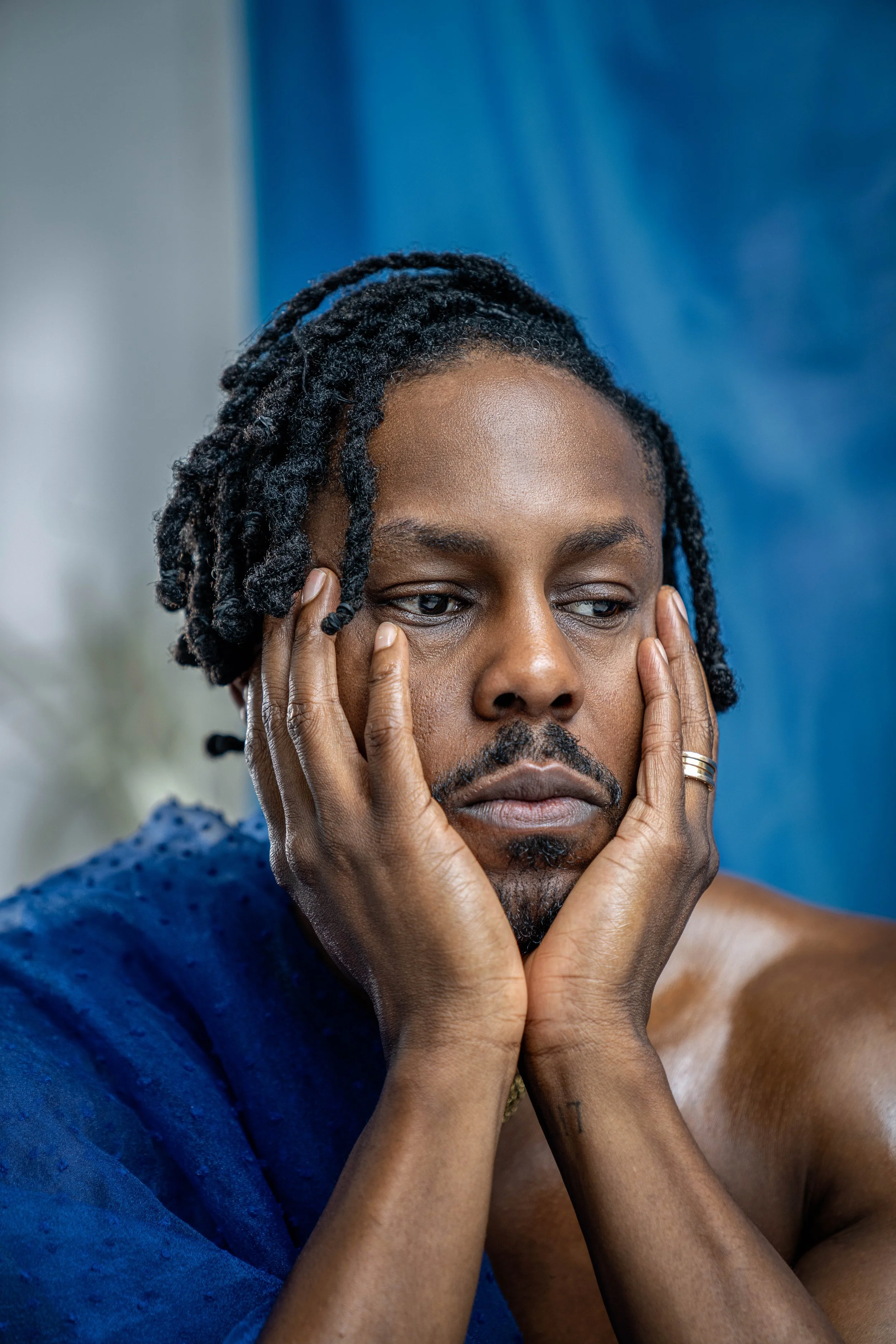 A person with dreadlocks and a mustache resting their face in their hands, looking contemplative, with a blue fabric background.