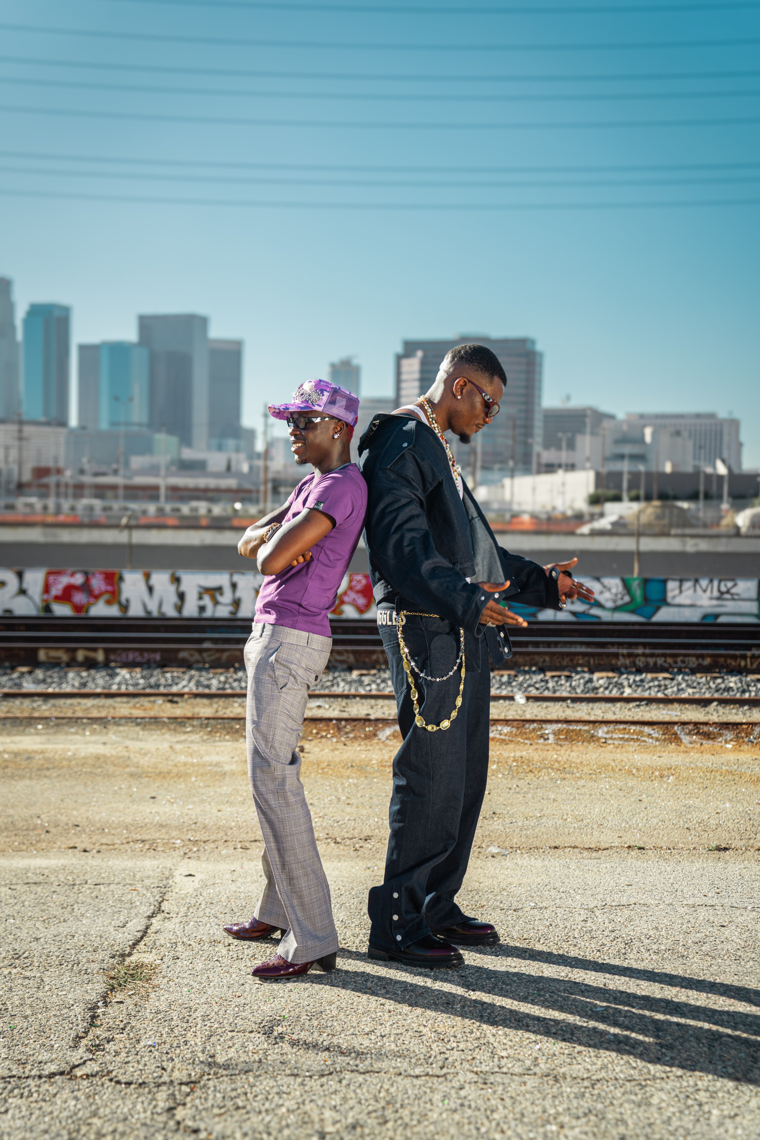 Two young men stand back-to-back on a train track in an urban area with a city skyline in the background. They are dressed in stylish, casual clothing and are looking at their phones, smiling and relaxed.