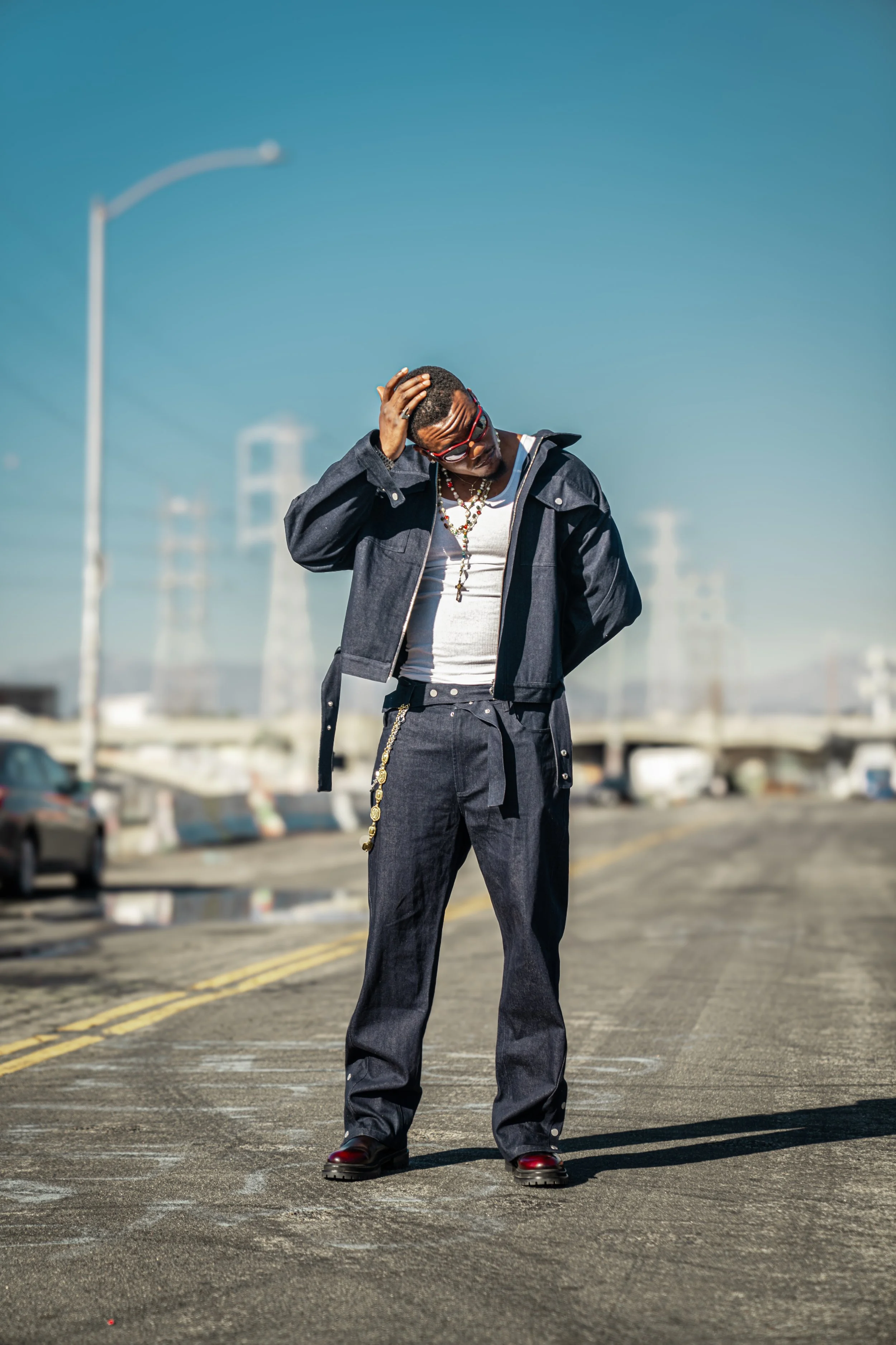 A man stands alone in the middle of a city street, wearing dark clothing, sunglasses, and jewelry, with a clear blue sky in the background.
