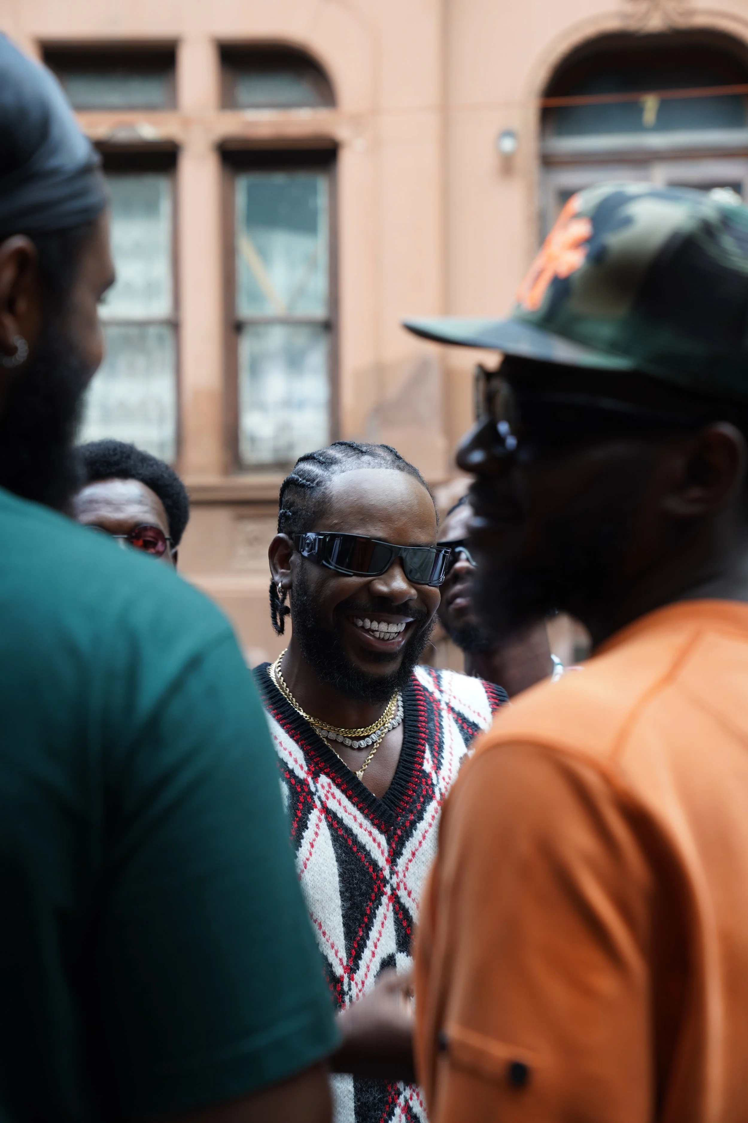 A group of people, with one man wearing sunglasses and a patterned sweater, smiling and engaging in conversation outdoors, with a building in the background.